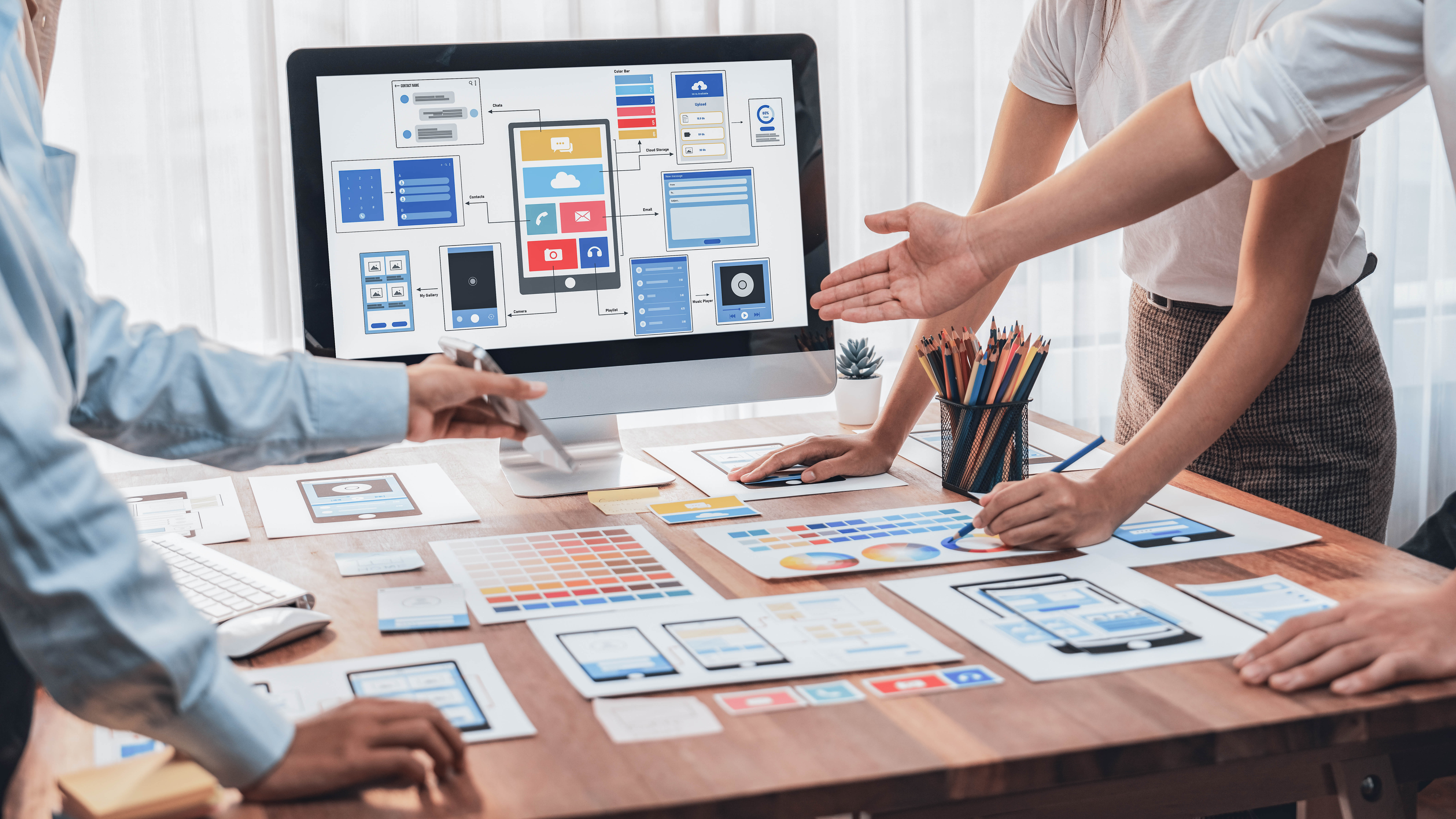 Mehrere Personen arbeiten an einem Tisch mit Designmaterialien, während ein Computer Bildschirm mit App-Layouts zeigt.