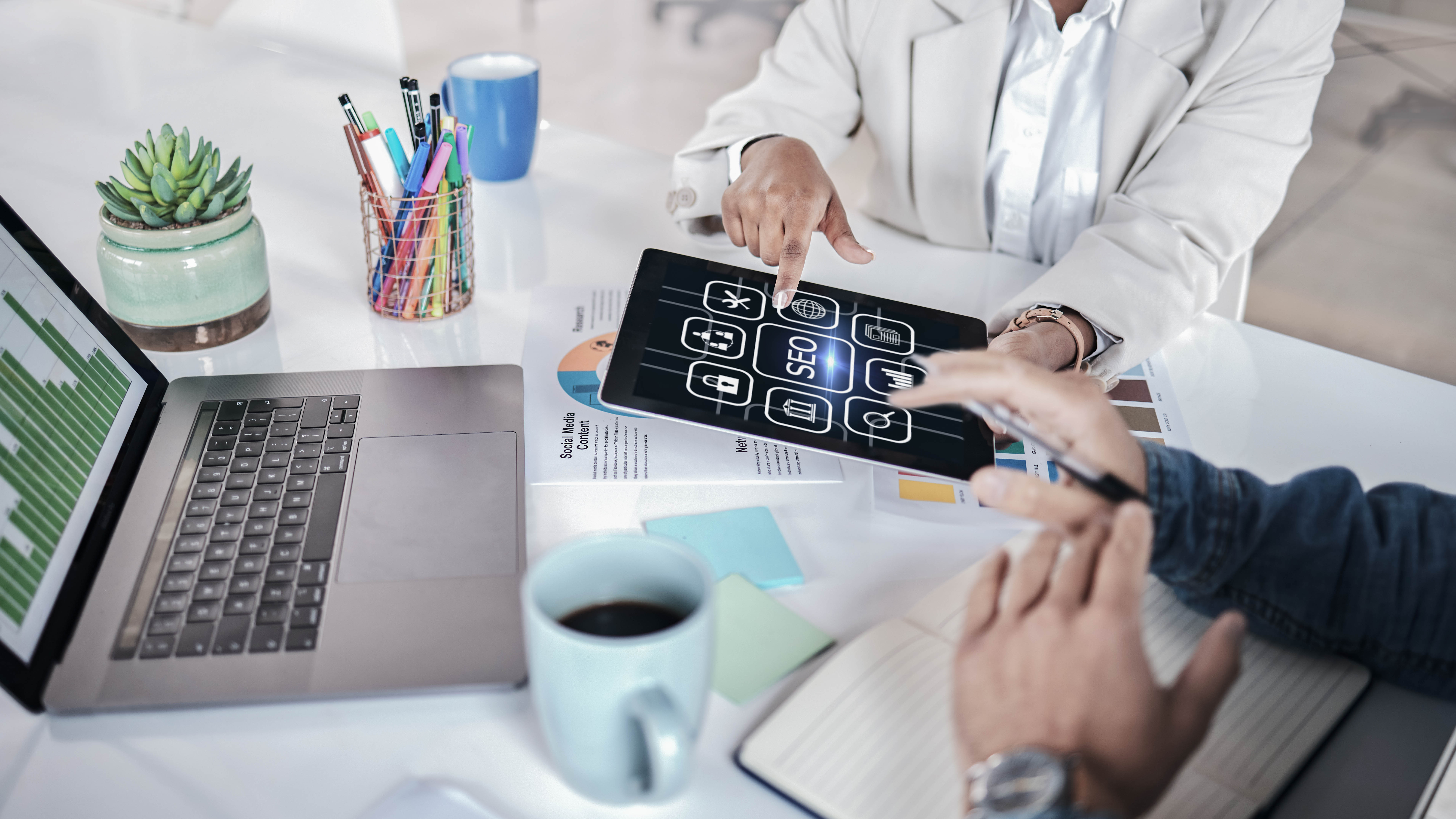 Zwei Personen diskutieren über digitale Strategien, während sie ein Tablet mit Icons und eine Laptop-Tastatur auf einem Tisch nutzen.