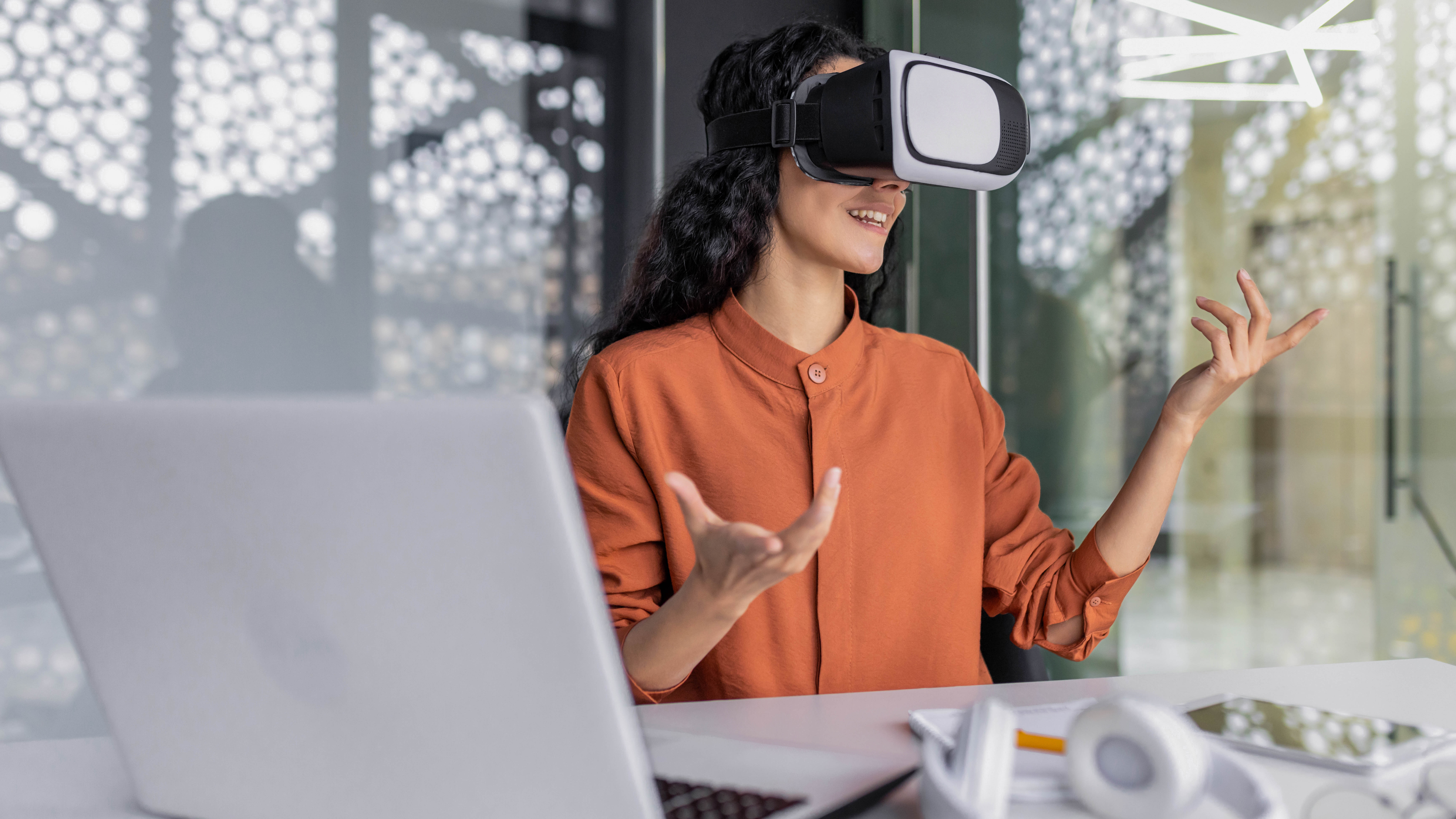 Frau mit VR-Brille sitzt an einem Tisch, gestikuliert und spricht, ein Laptop und Kopfhörer sind sichtbar.