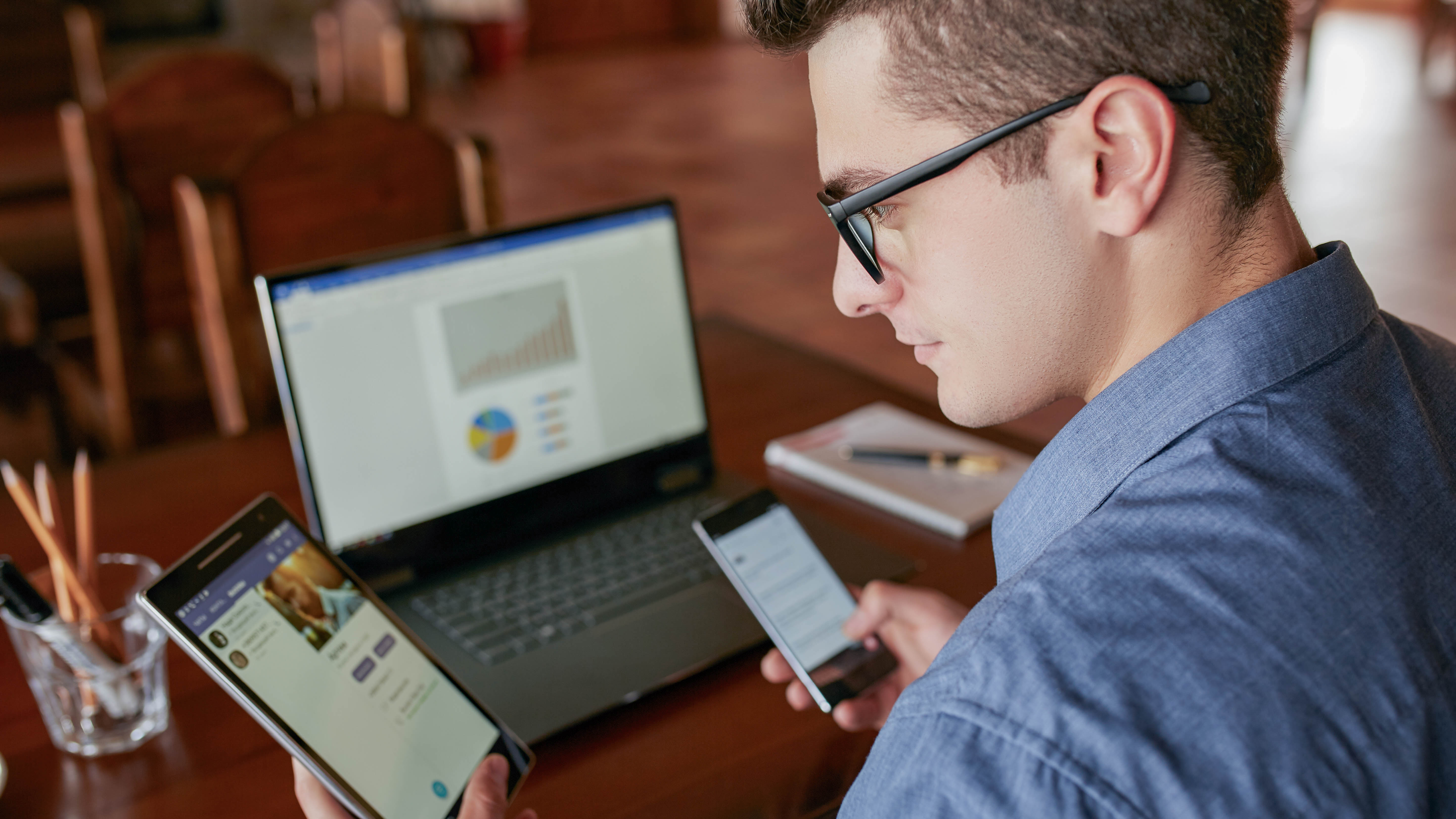 Mann mit Brille nutzt ein Tablet und ein Smartphone, während ein Laptop mit Diagrammen im Hintergrund steht.