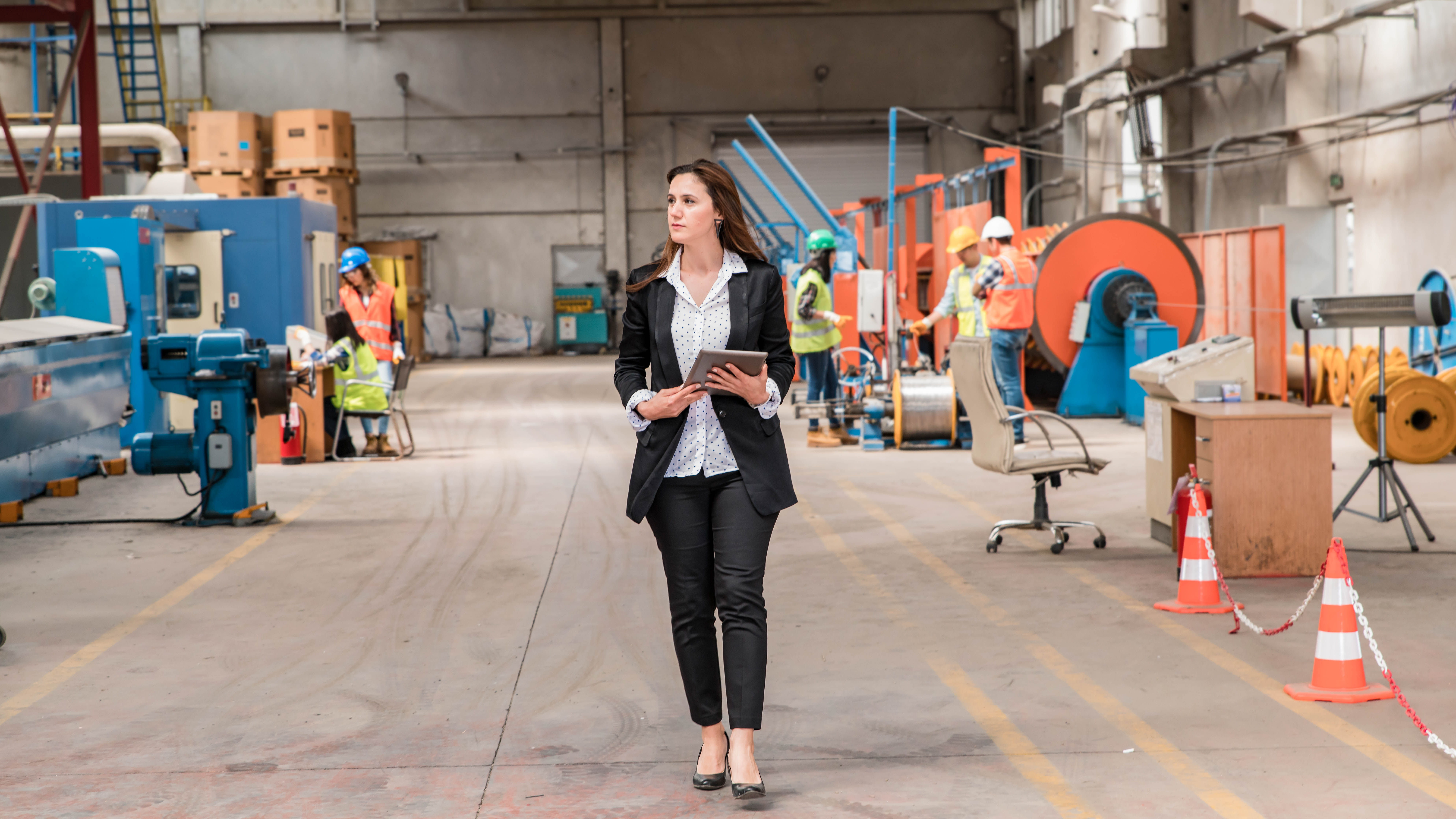 Eine Frau in einem Anzug mit Tablet geht durch eine Industriehalle, umgeben von Arbeitern in Schutzkleidung.