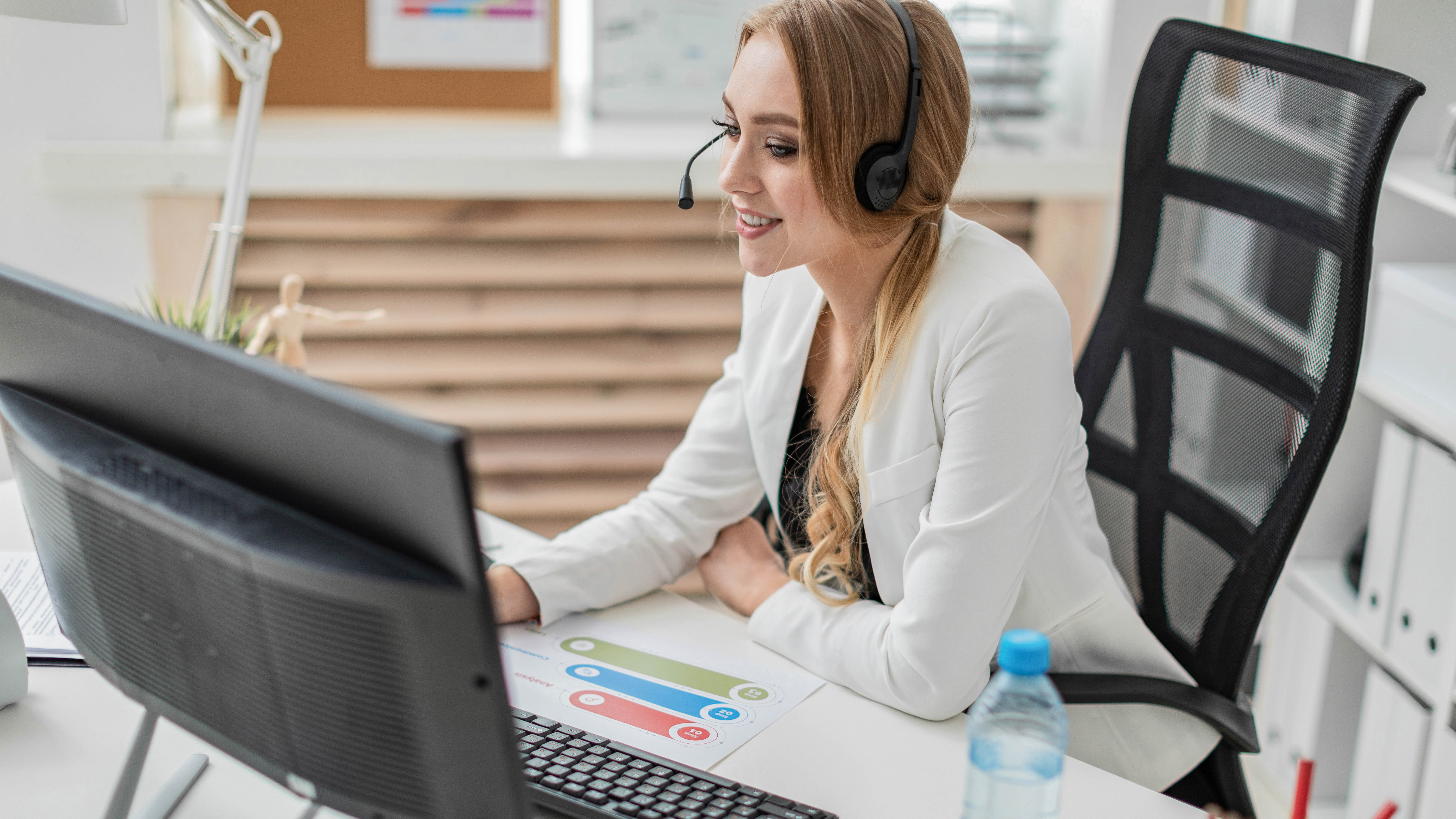 Frau mit Headset sitzt an einem Schreibtisch und arbeitet am Computer, neben ihr steht eine Wasserflasche.
