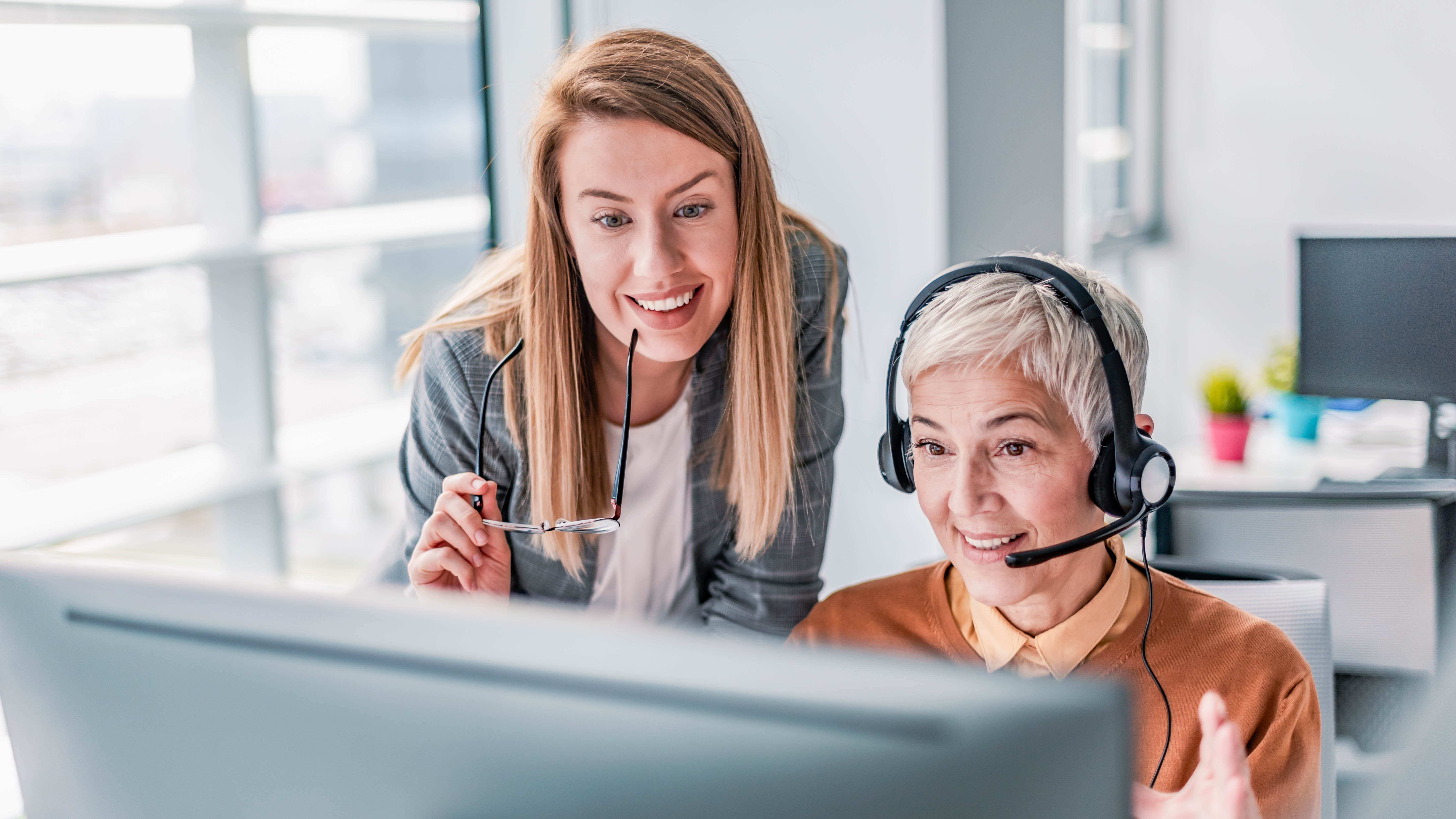 Zwei Frauen in einem modernen Büro schauen gemeinsam auf einen Computerbildschirm, eine trägt ein Headset.