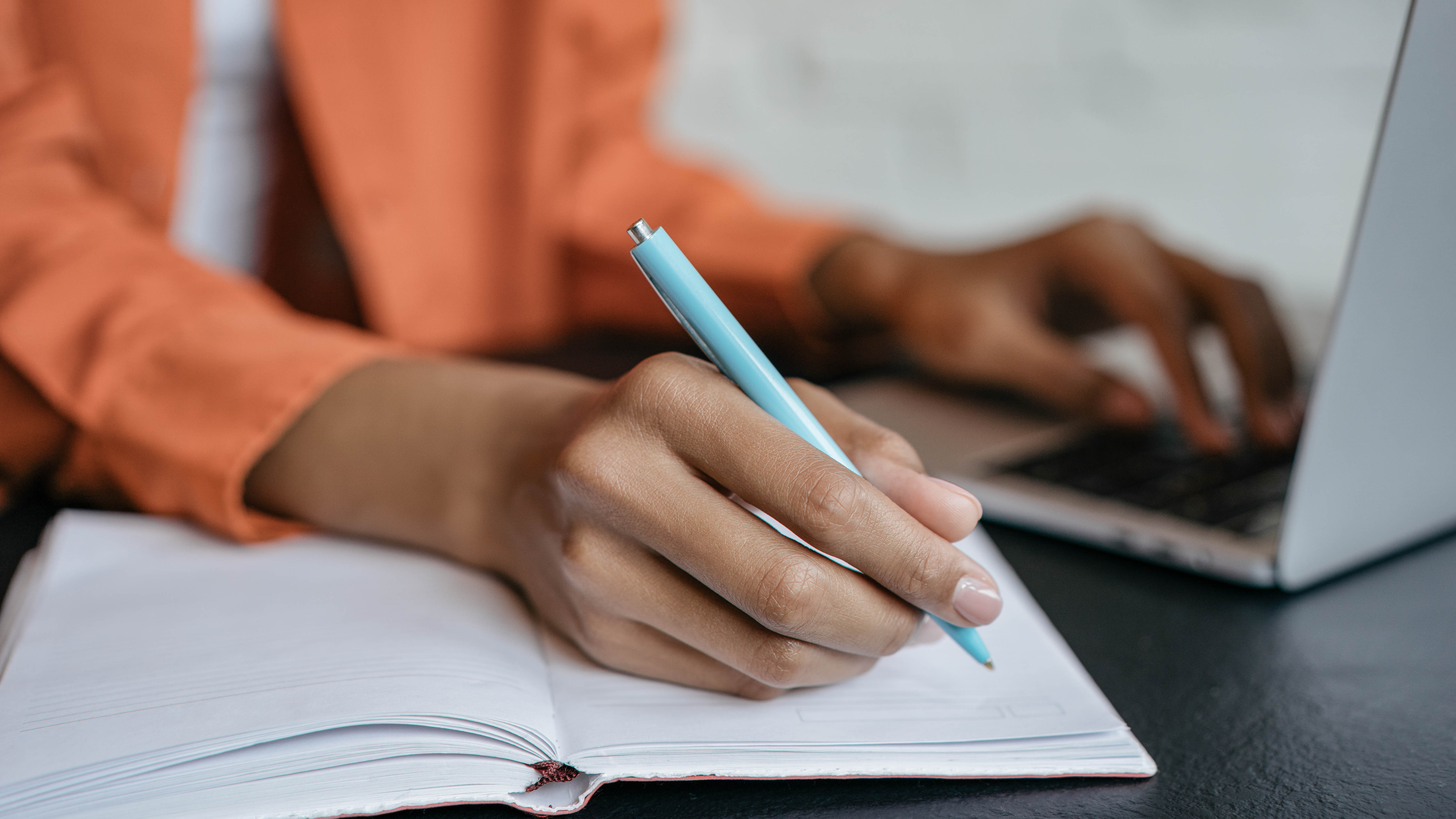 Eine Hand mit einem blauen Stift schreibt in ein offenes Notizbuch, während eine andere Hand auf einem Laptop tippt.