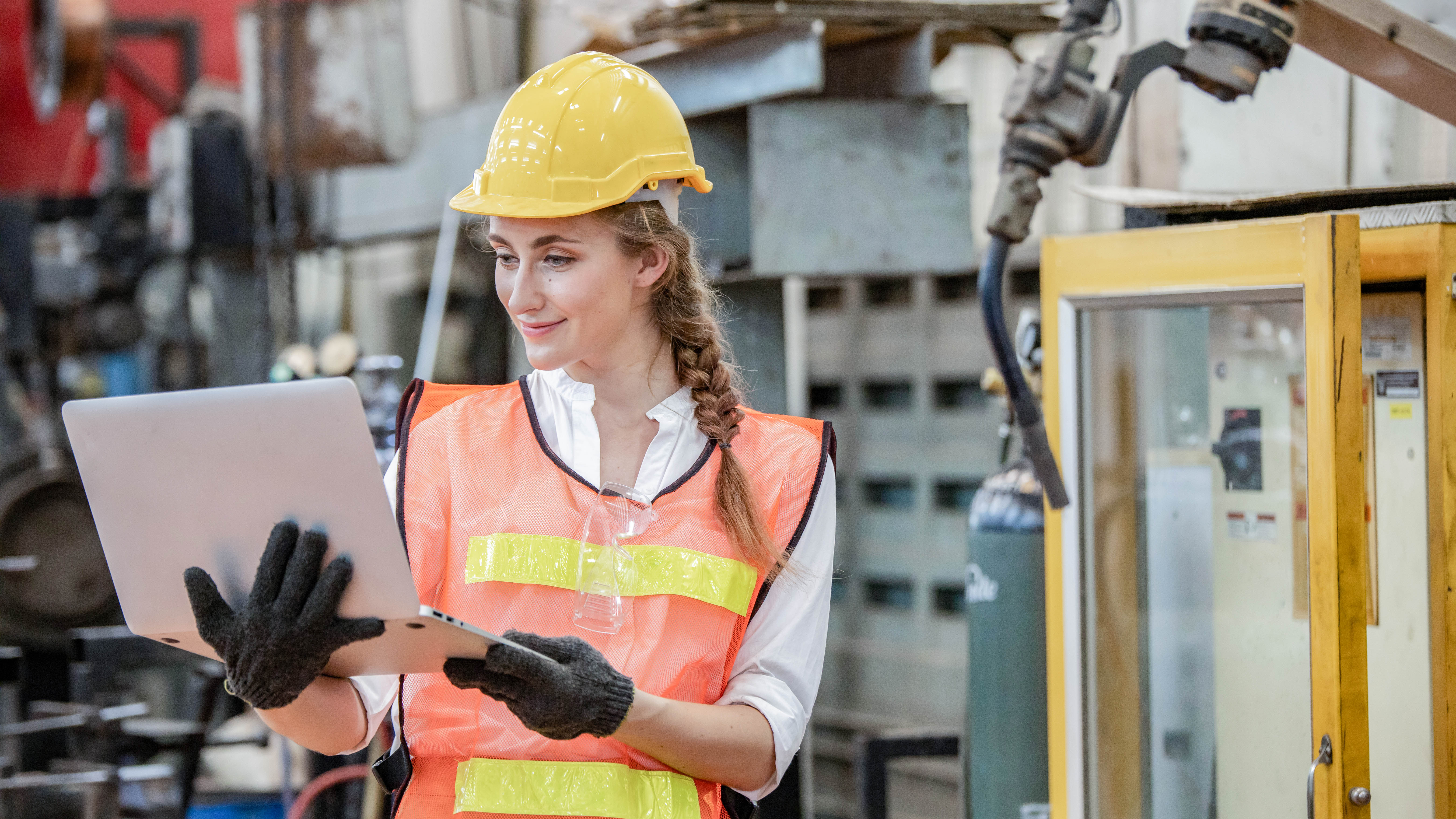 Frau in Sicherheitskleidung und Helm arbeitet mit einem Laptop in einer Industrieumgebung.