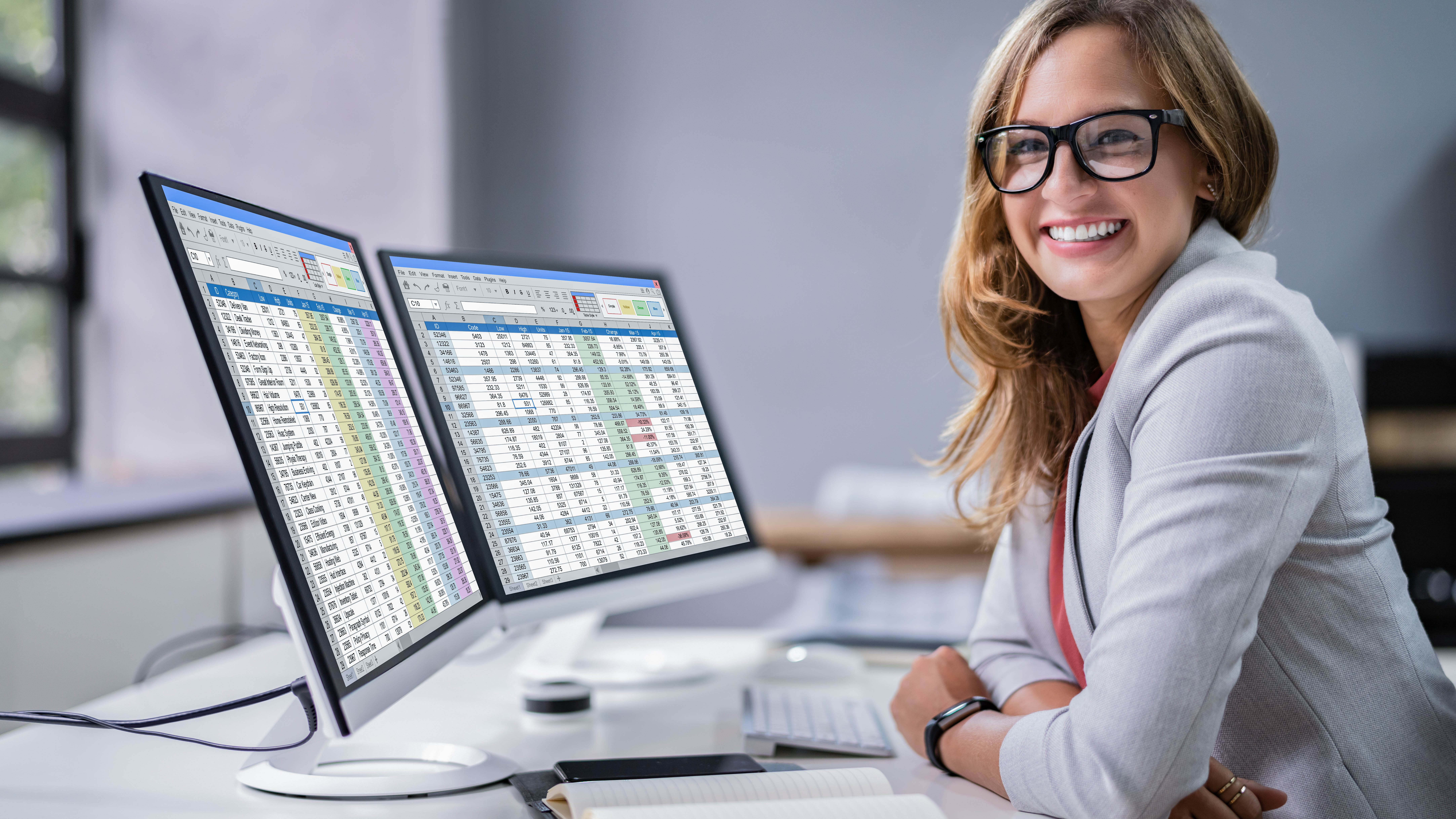 Frau mit Brille lächelt am Schreibtisch, zwei Monitore zeigen Tabellenkalkulationen, Notizbuch und Tastatur sichtbar.