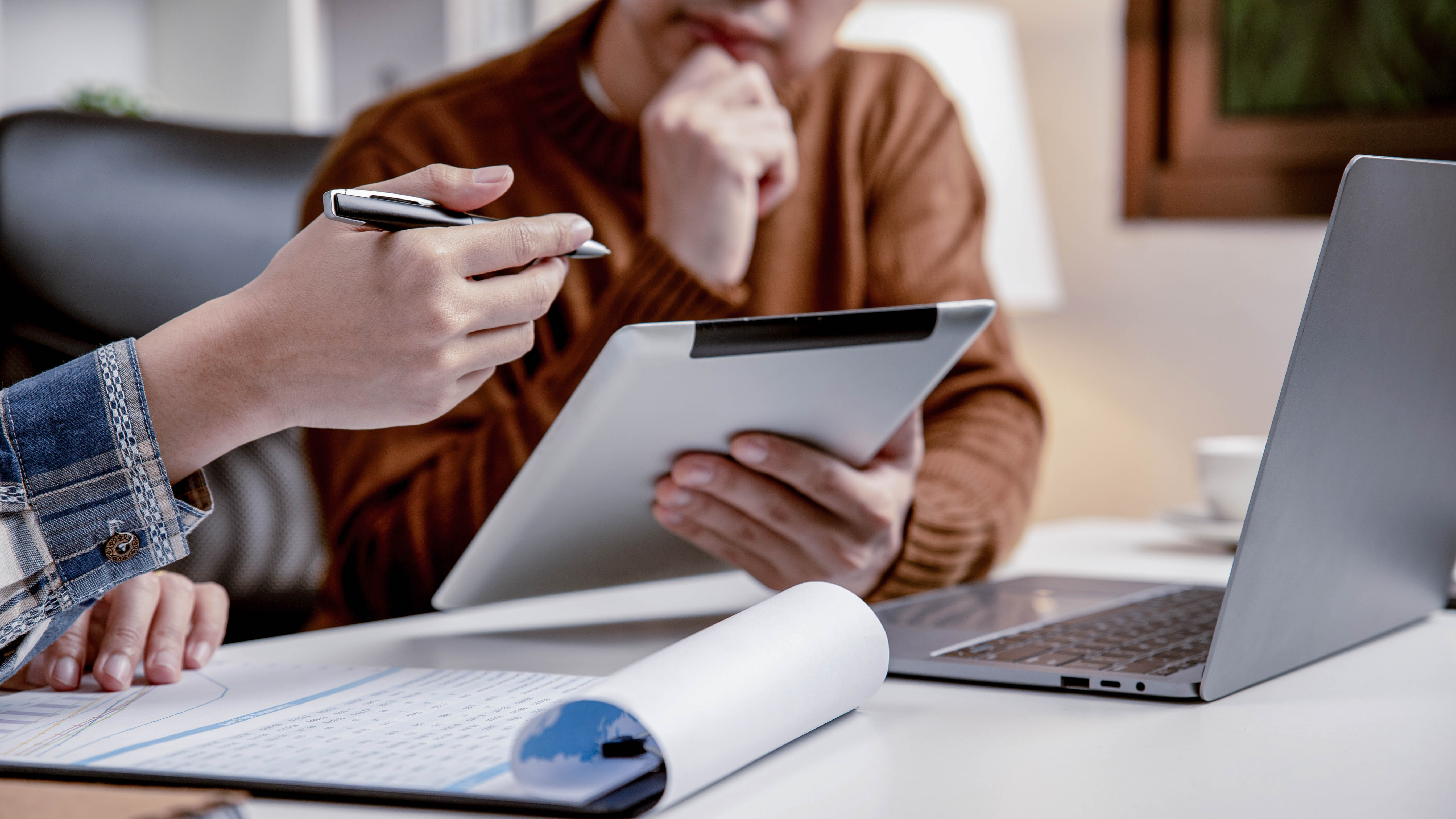 Hände halten ein Tablet und einen Stift, während ein Laptop und ein aufgerolltes Dokument auf einem Tisch liegen.