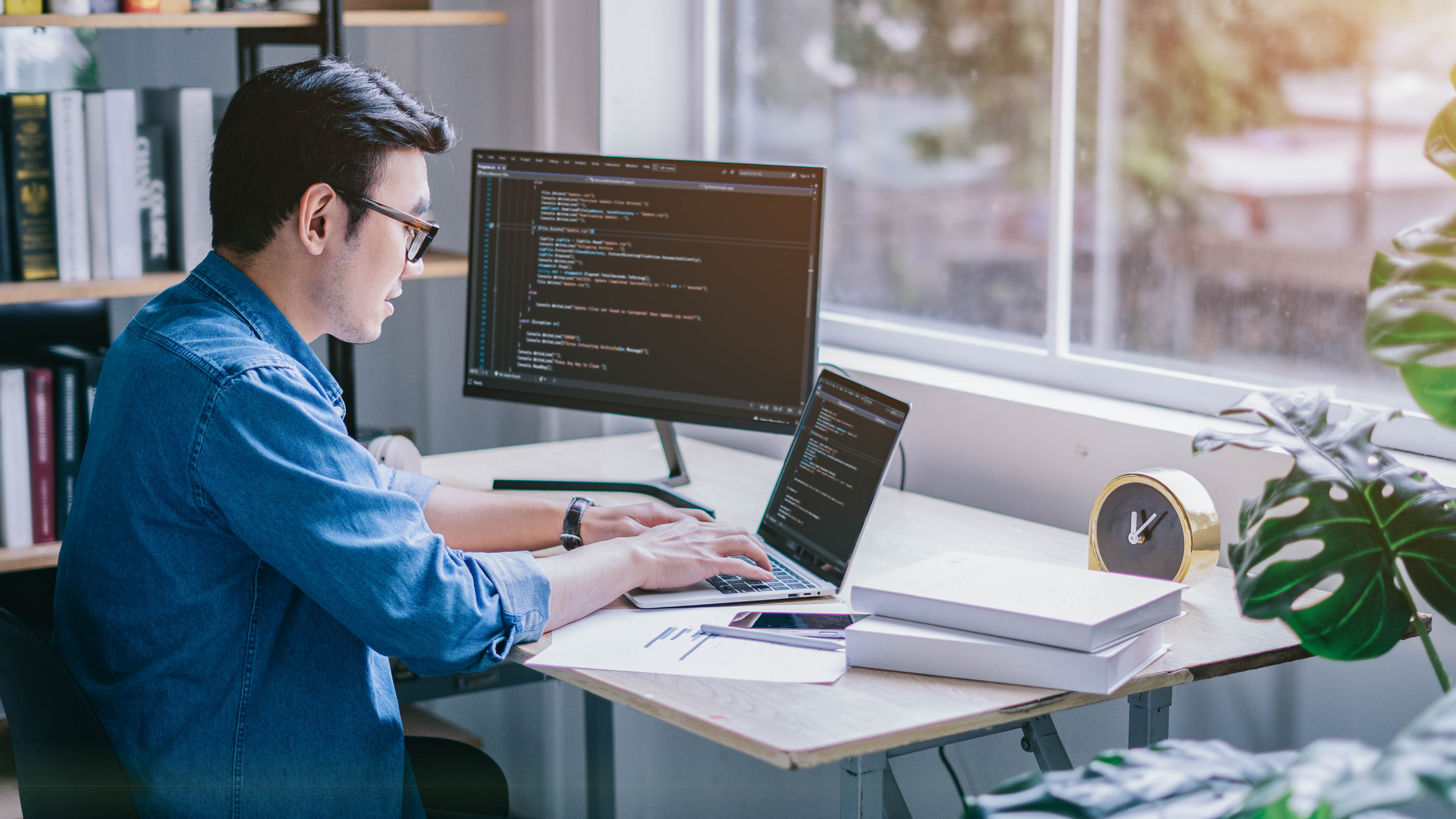 Mann arbeitet an einem Laptop und einem Monitor mit Programmiercode, umgeben von Pflanzen und Büchern.