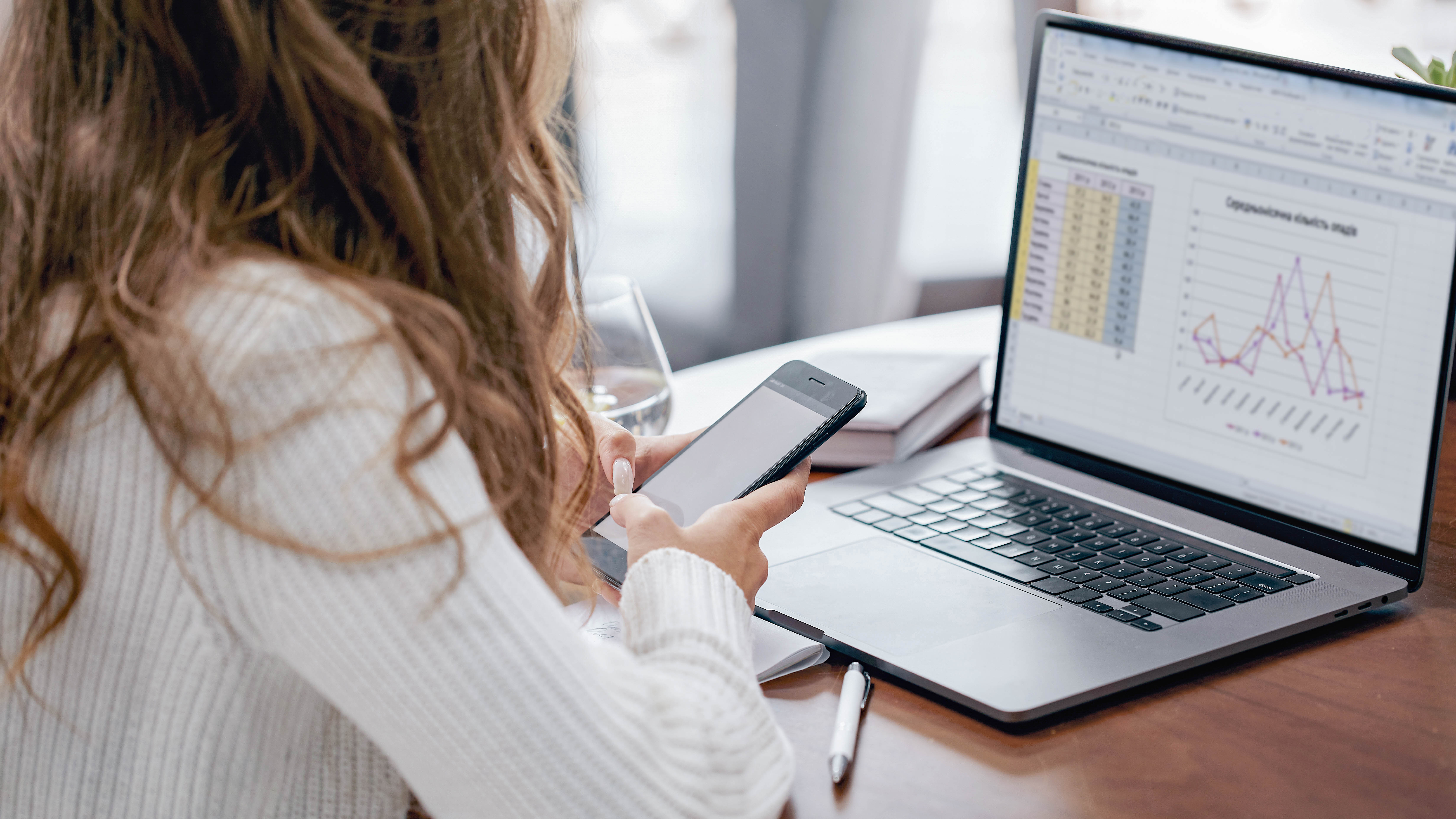 Eine Person mit lockigem Haar sitzt an einem Tisch und nutzt ein Smartphone, während ein Laptop mit Daten und Diagrammen geöffnet ist.