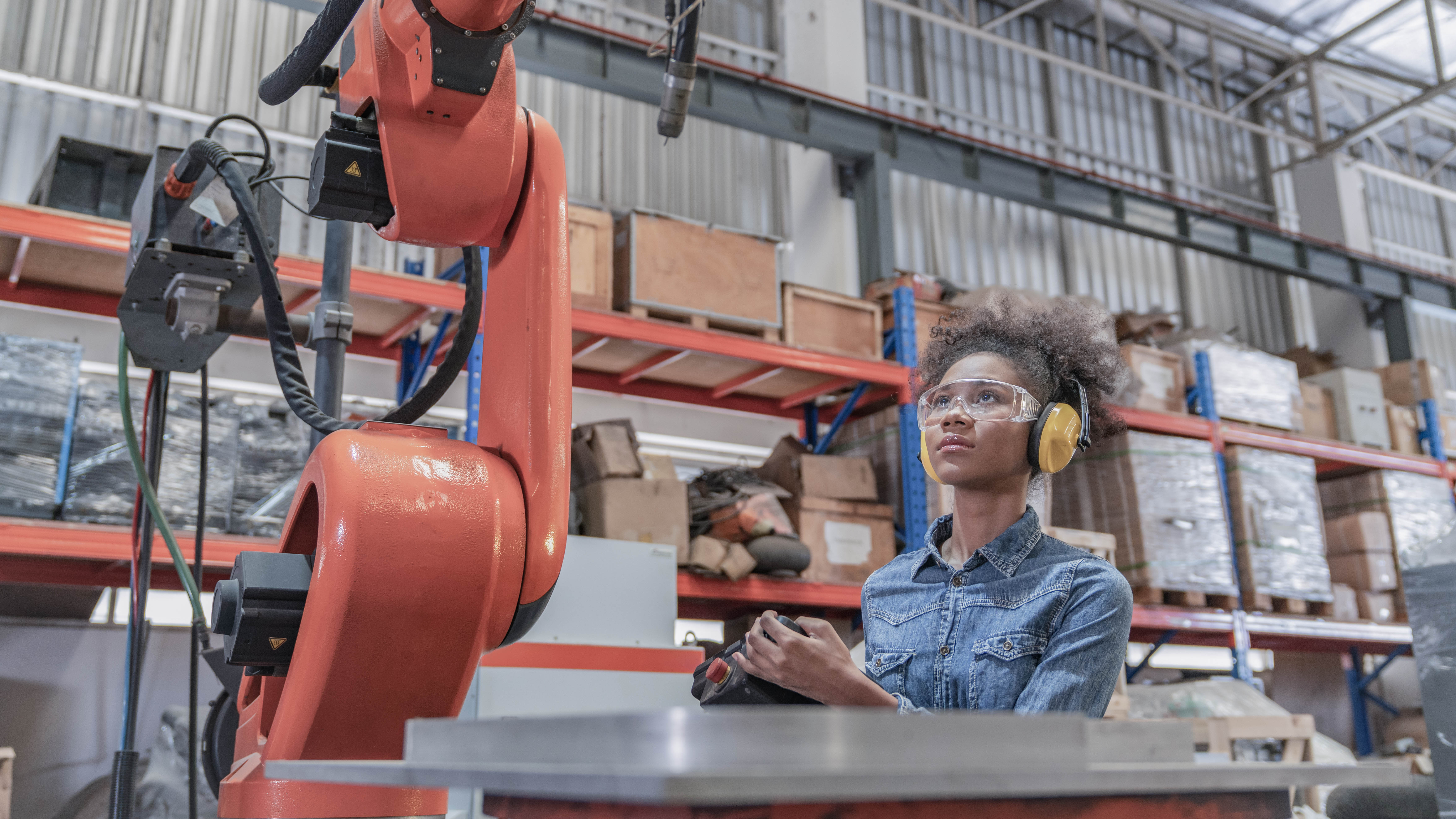 Eine Person mit Schutzbrille und Ohrenschützern beobachtet einen Industrieroboter in einer Lagerhalle.