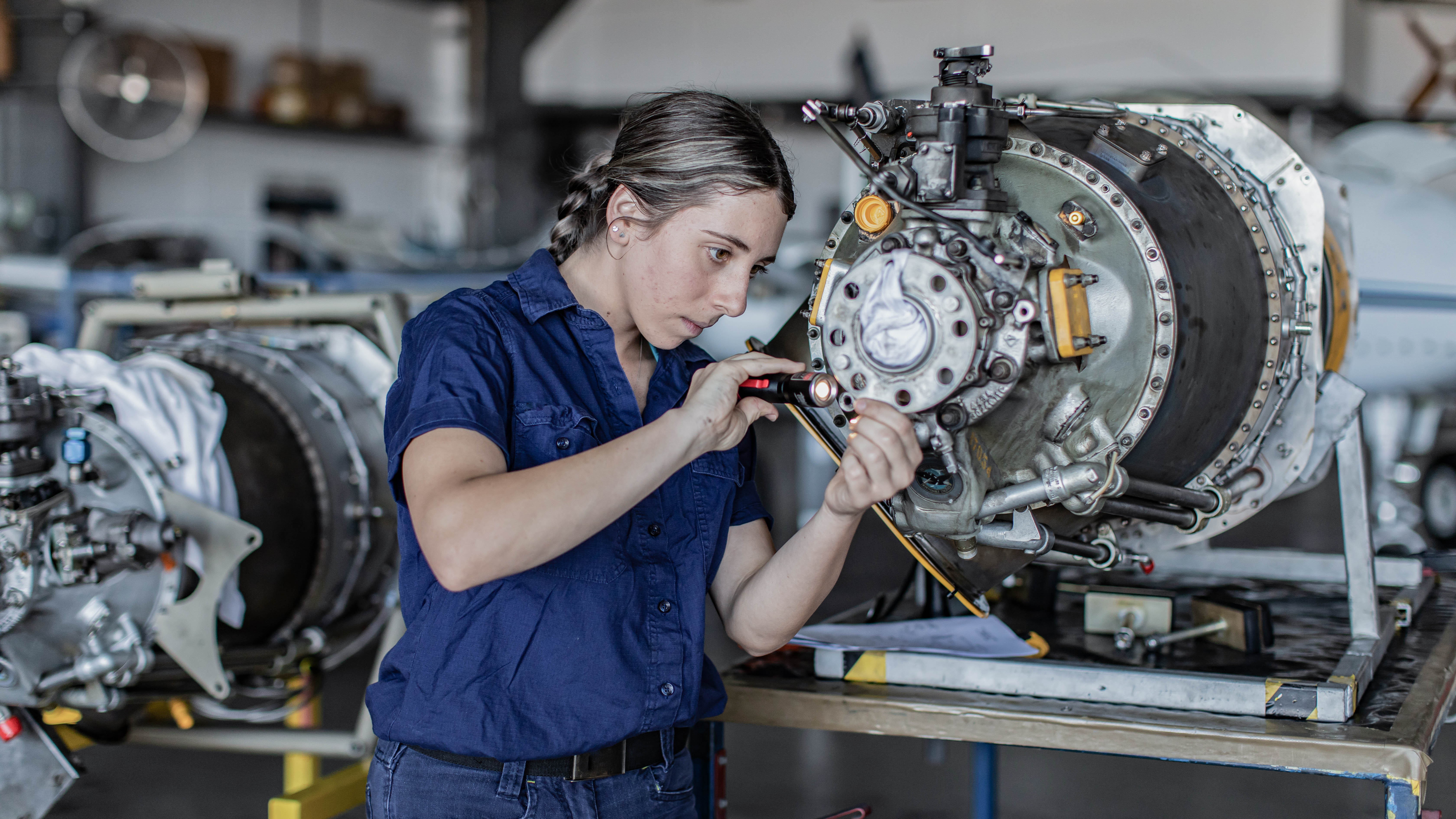 Eine Mechanikerin arbeitet konzentriert an einem Flugzeugmotor und beleuchtet ihn mit einer Taschenlampe.
