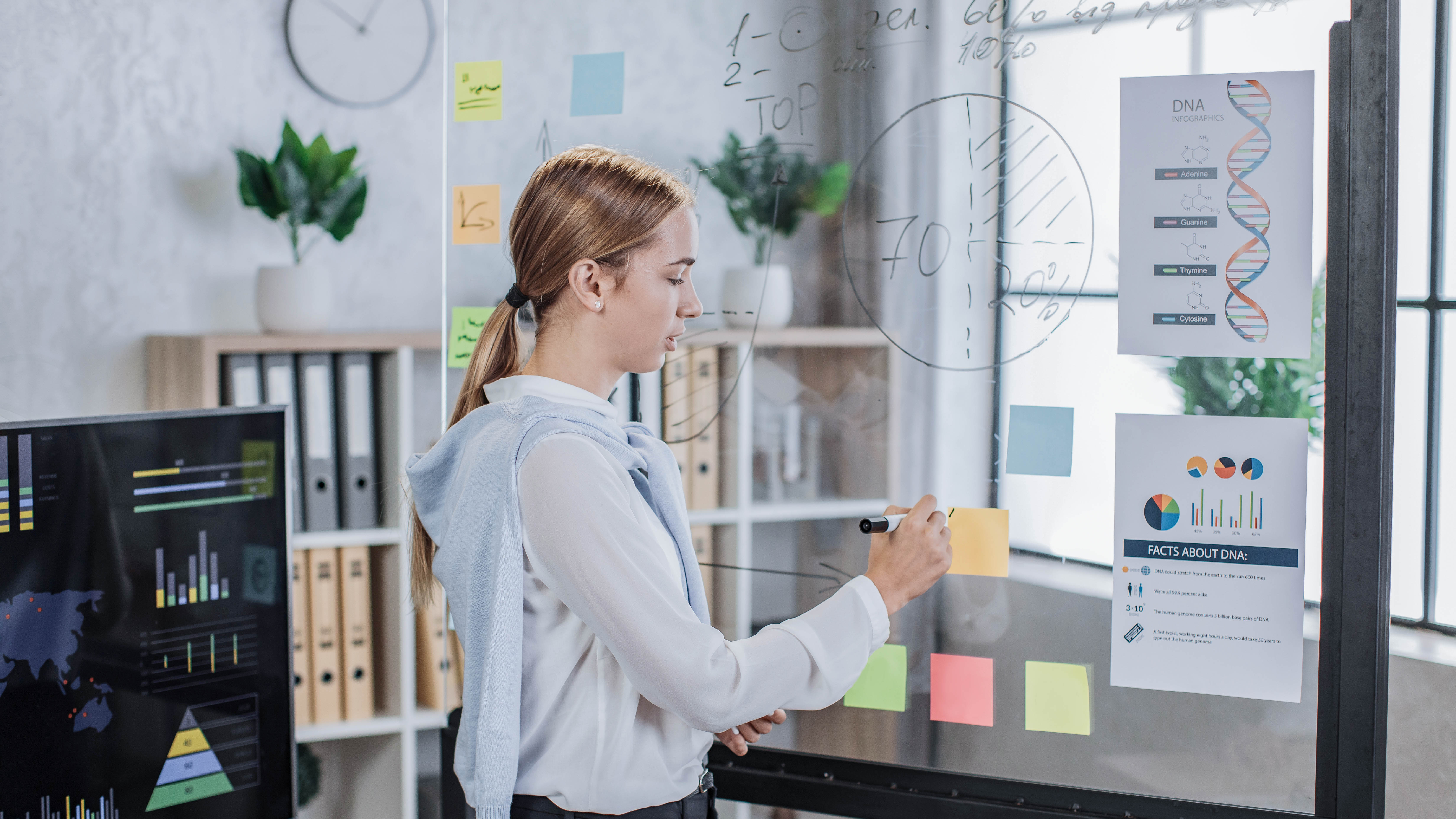 Eine Person schreibt mit einem Marker auf eine transparente Tafel, umgeben von bunten Haftnotizen und Informationsblättern.