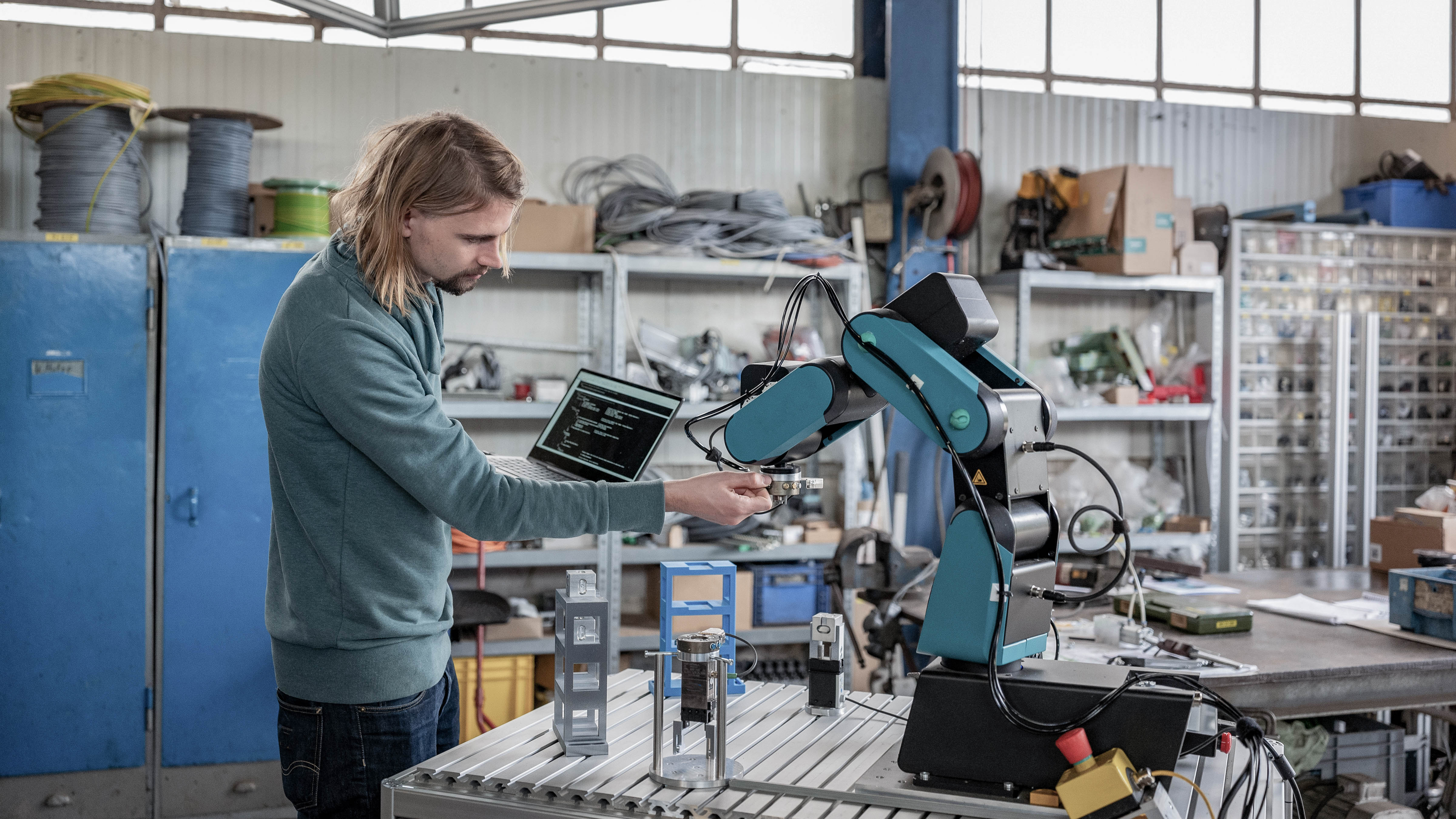 Ein Mann arbeitet an einem Roboterarm und hält ein Bauteil, während ein Laptop auf dem Tisch steht.