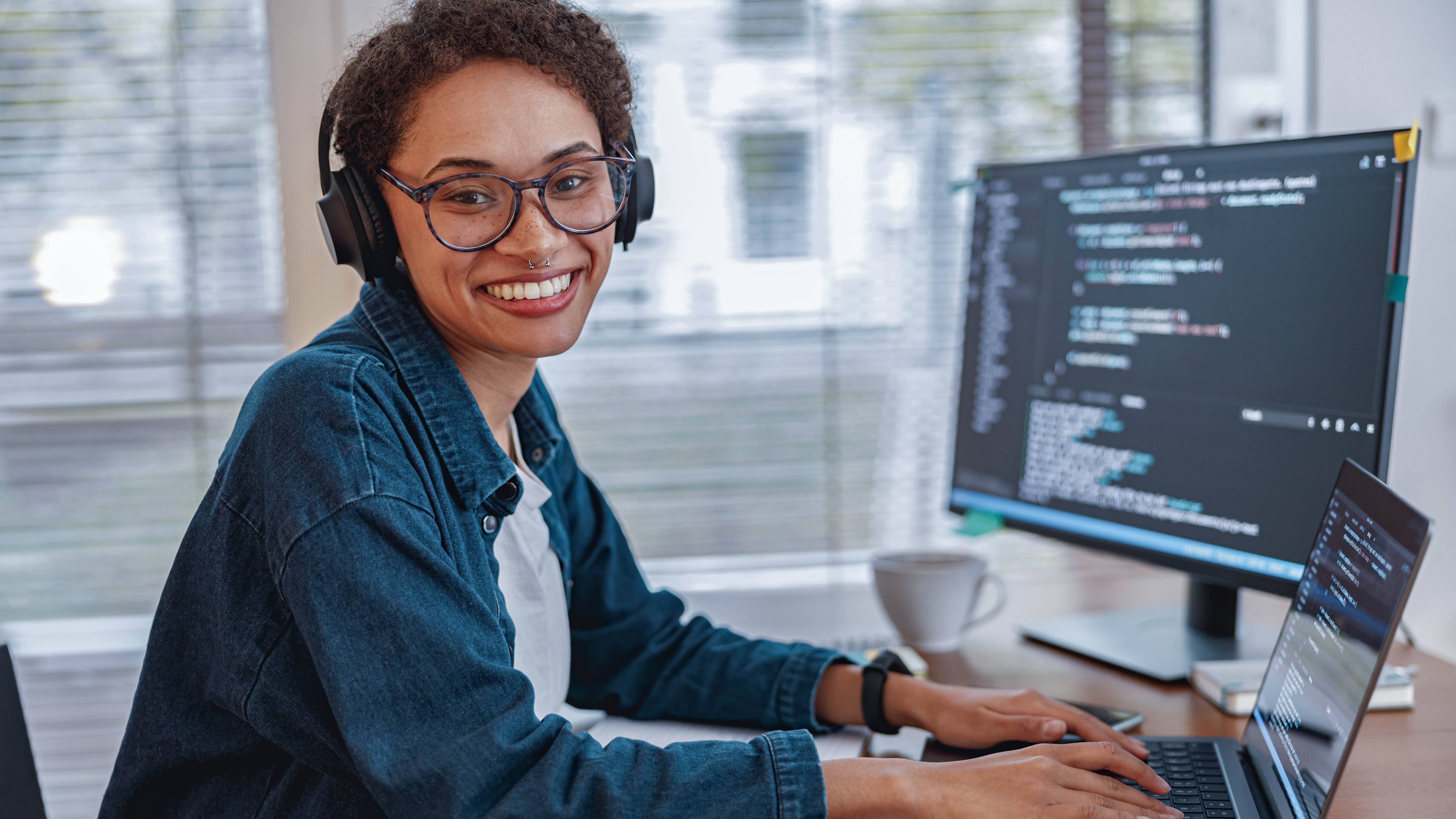 Lächelnde Person mit Kopfhörern arbeitet an einem Laptop und einem Monitor mit Programmiercode.