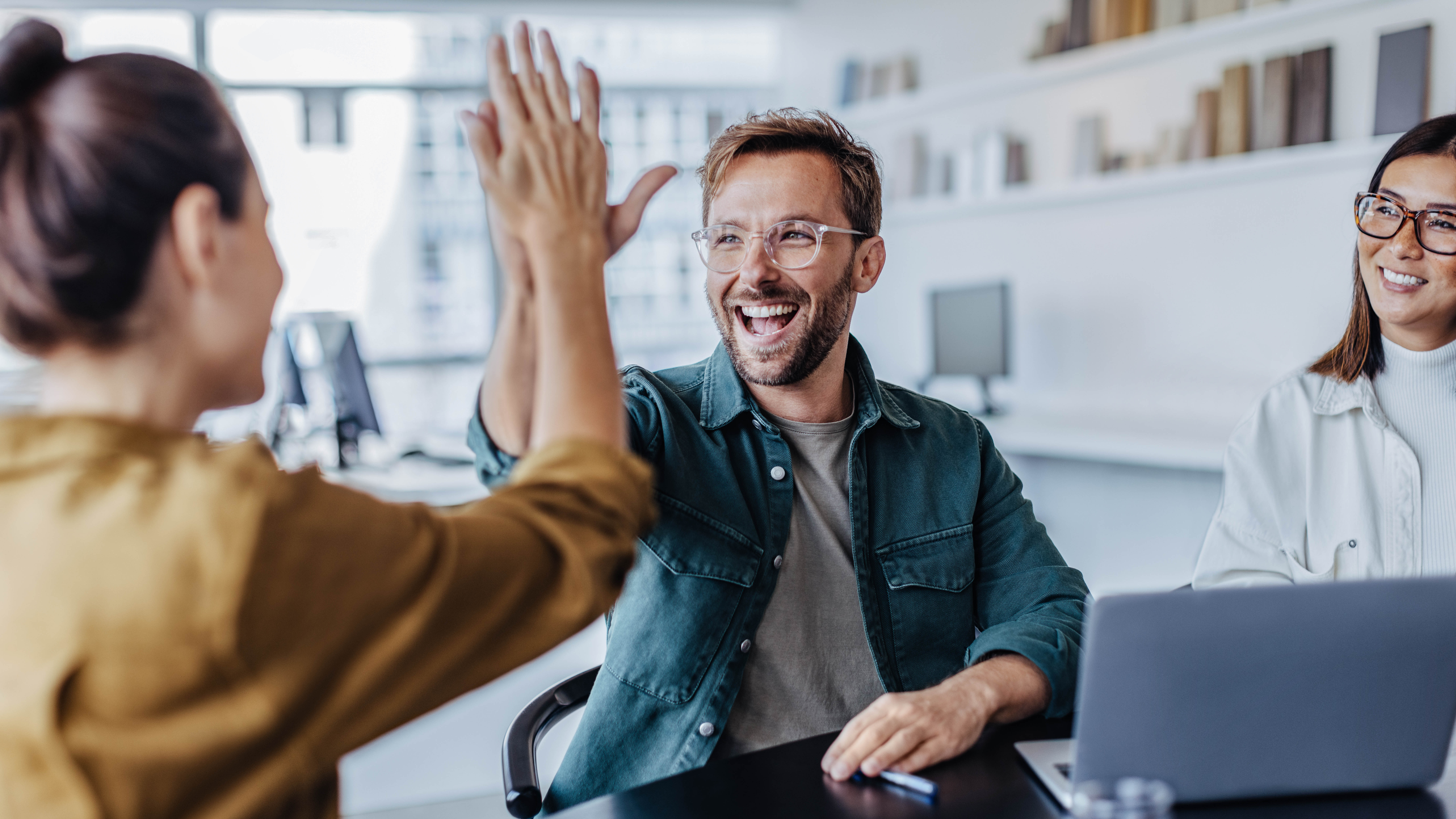 Ein Mann und eine Frau geben sich in einem modernen Büro ein High-Five, während eine weitere Frau lächelt.