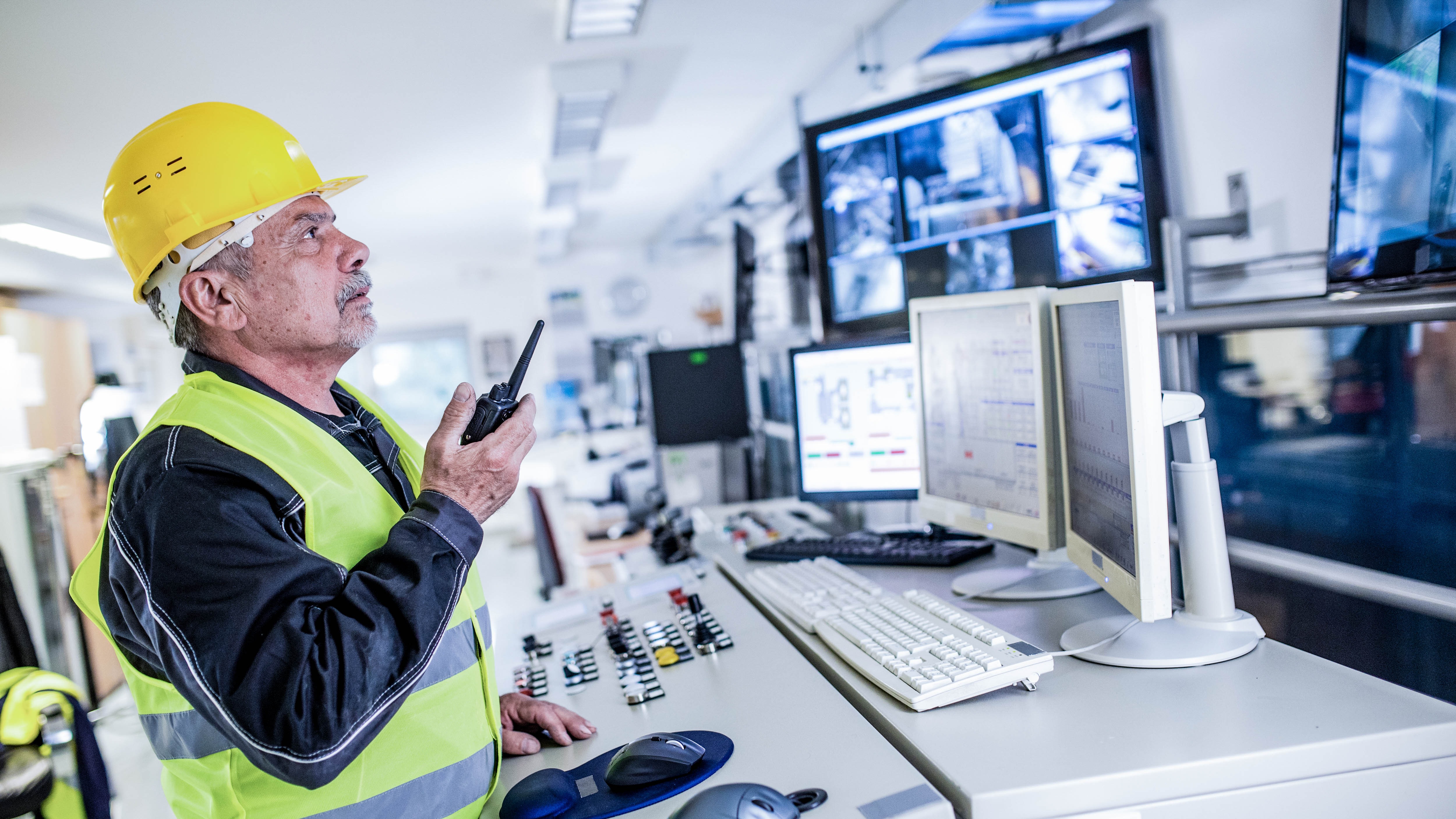Ein Mann mit gelbem Helm und Warnweste spricht mit einem Funkgerät vor mehreren Monitoren in einem Kontrollraum.