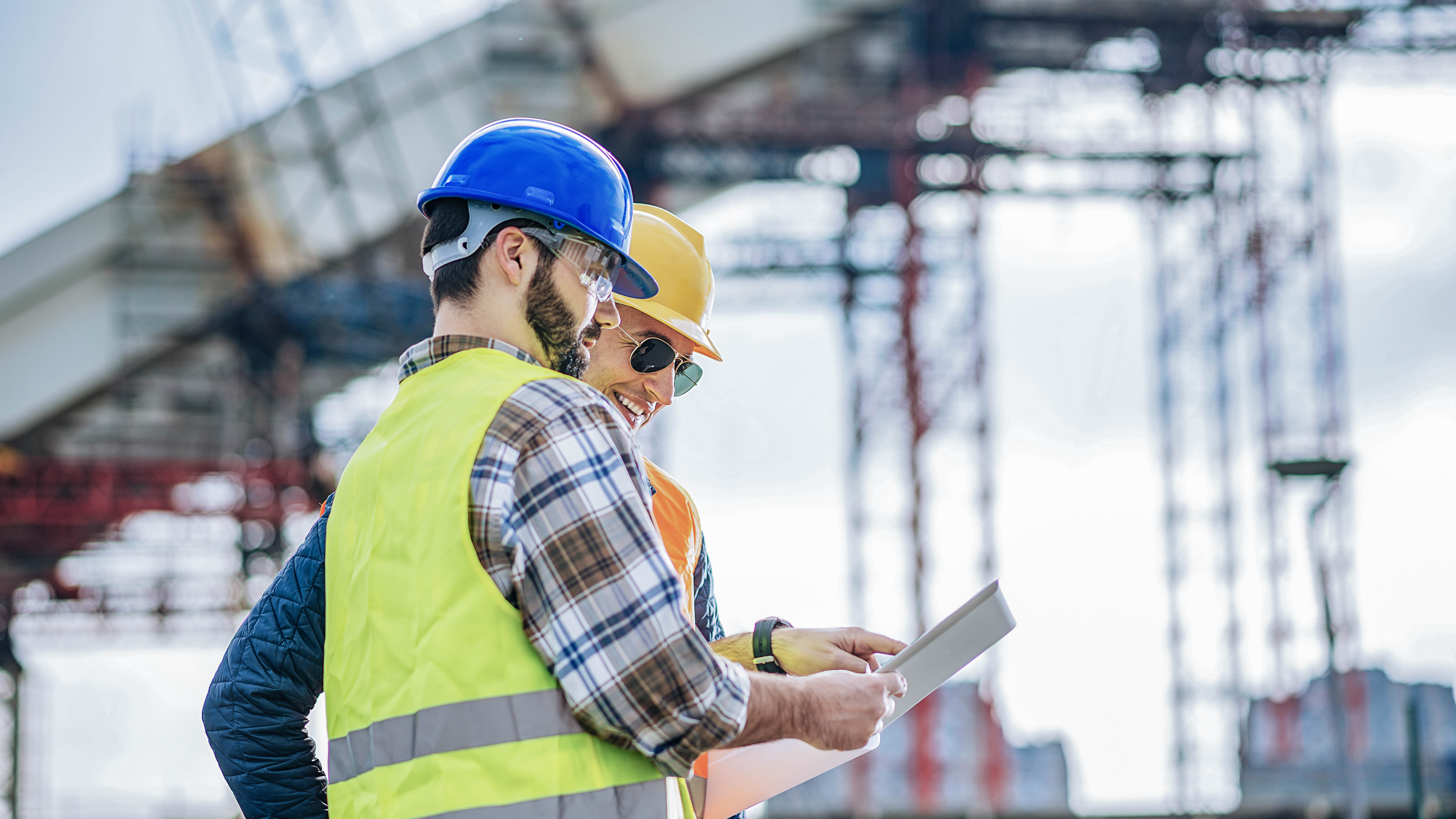 Zwei Bauarbeiter in Schutzhelmen und Warnwesten betrachten Pläne auf einer Baustelle.