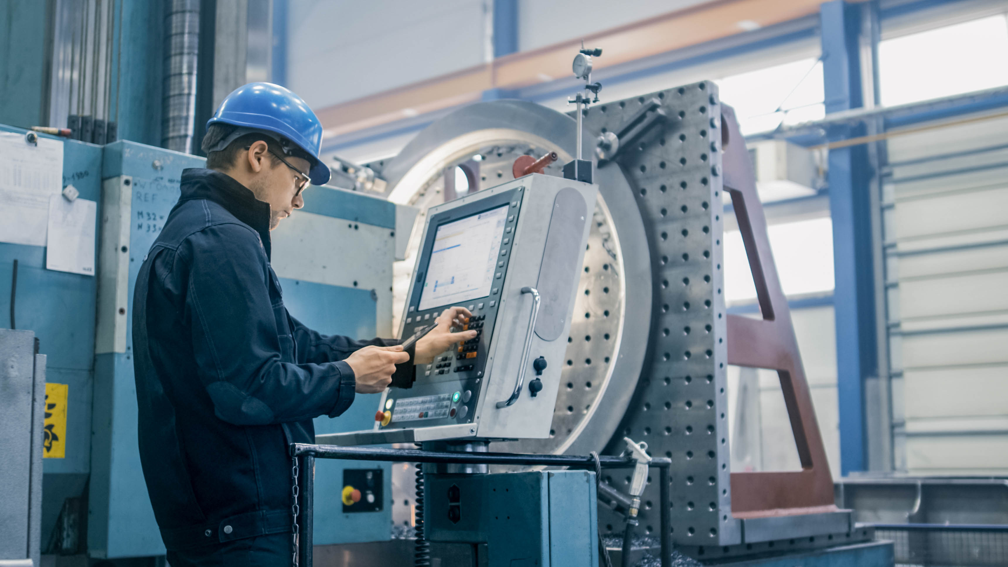 Ein Arbeiter in einem blauen Helm bedient eine CNC-Maschine in einer industriellen Umgebung.