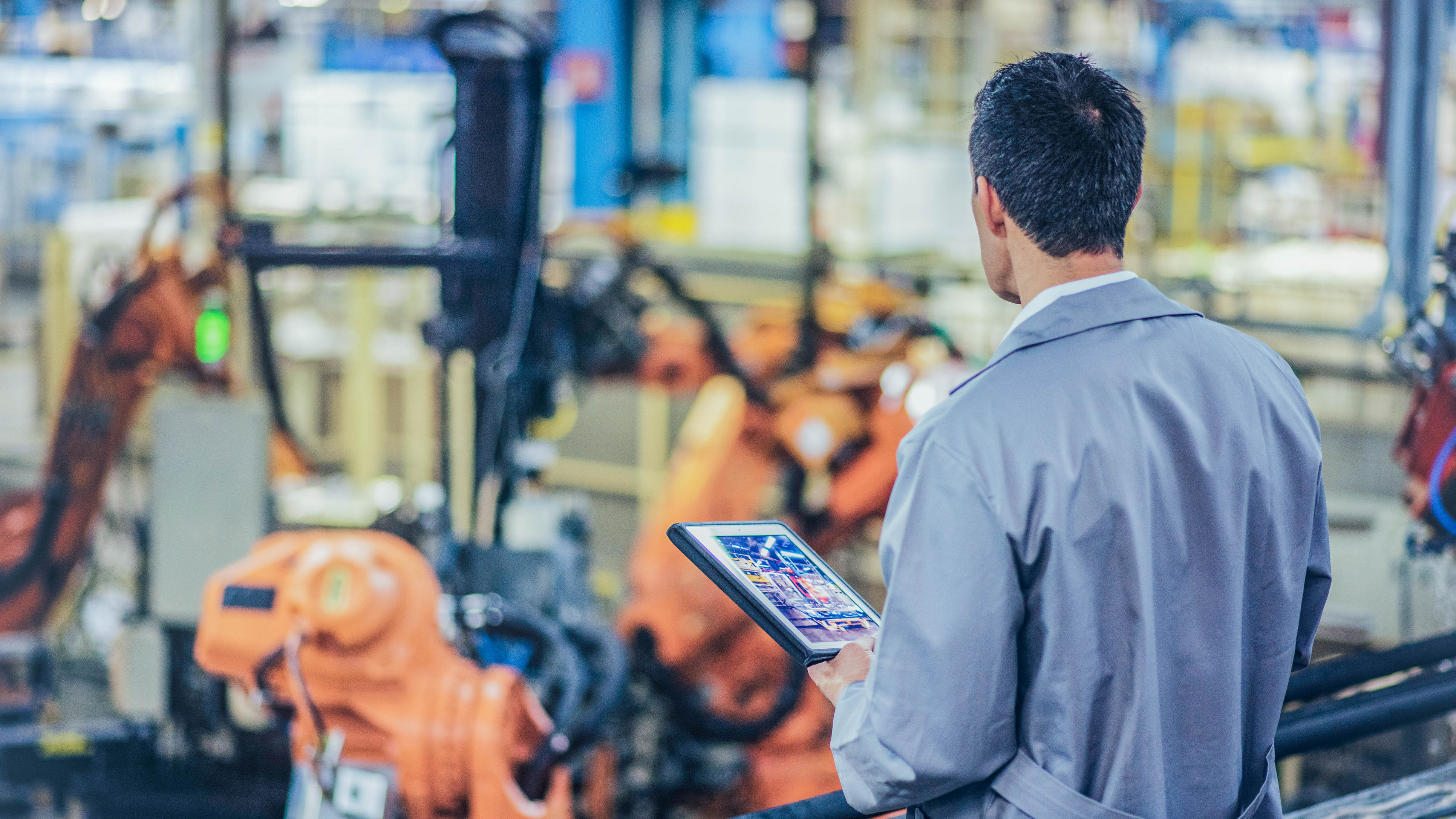 Ein Mann in einem grauen Arbeitsmantel steht mit einem Tablet in der Hand vor industriellen Robotern in einer Fabrik.