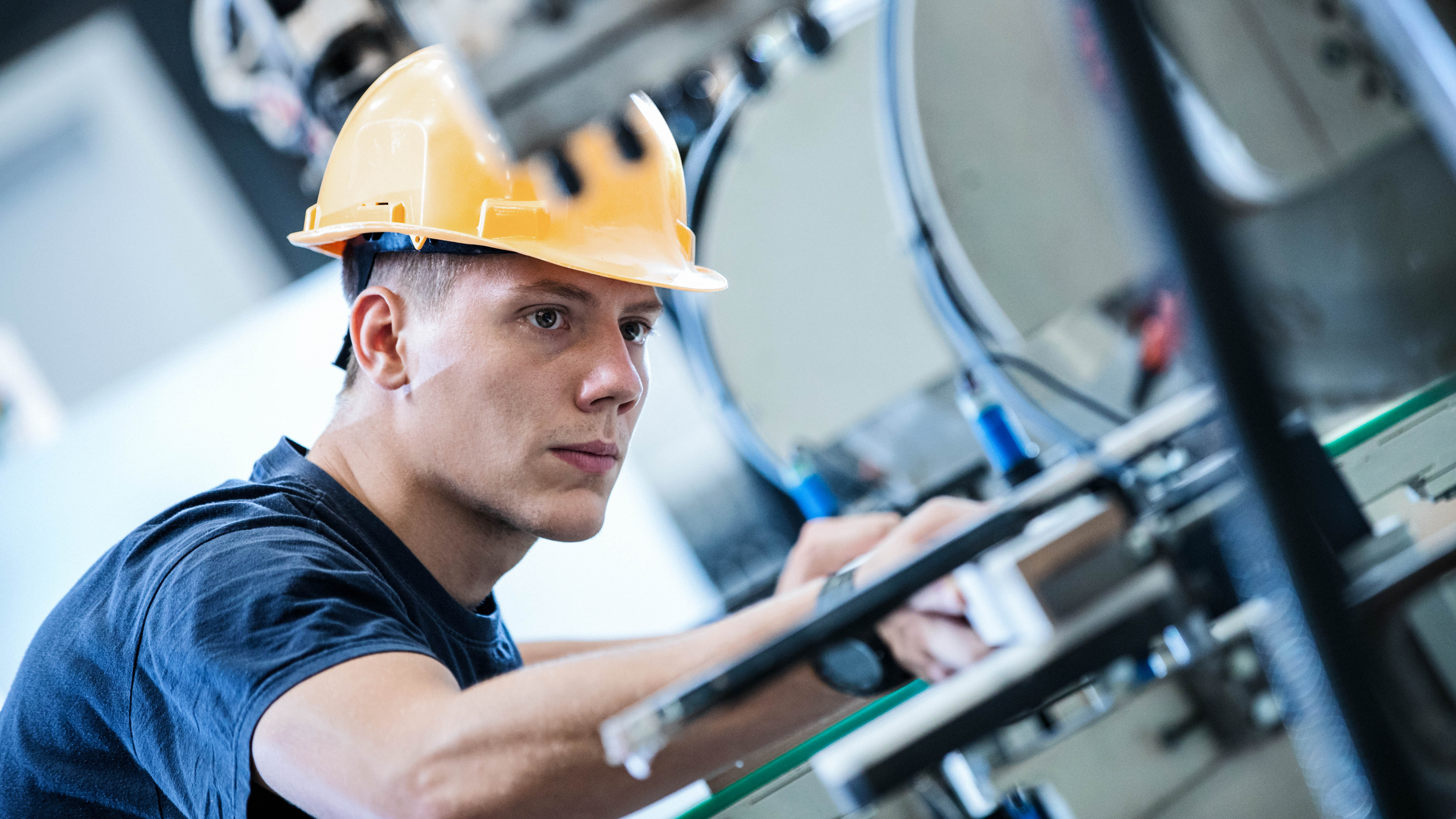 Junger Mann mit gelbem Helm arbeitet konzentriert an einer Maschine in einer Werkstatt.