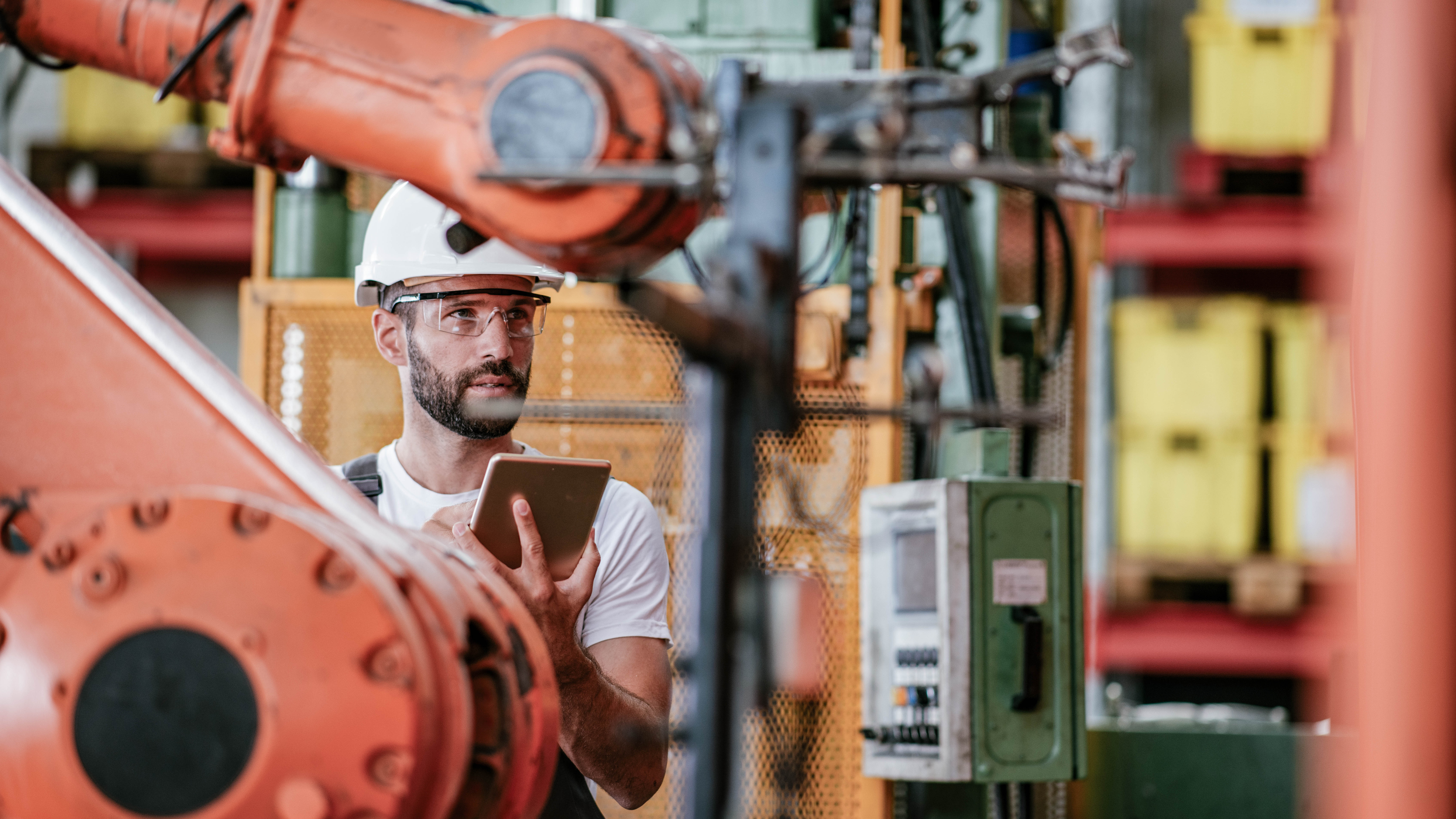 Ein Mann mit Schutzhelm und Brille steht in einer Fabrik und hält ein Tablet, während er einen Industrieroboter betrachtet.