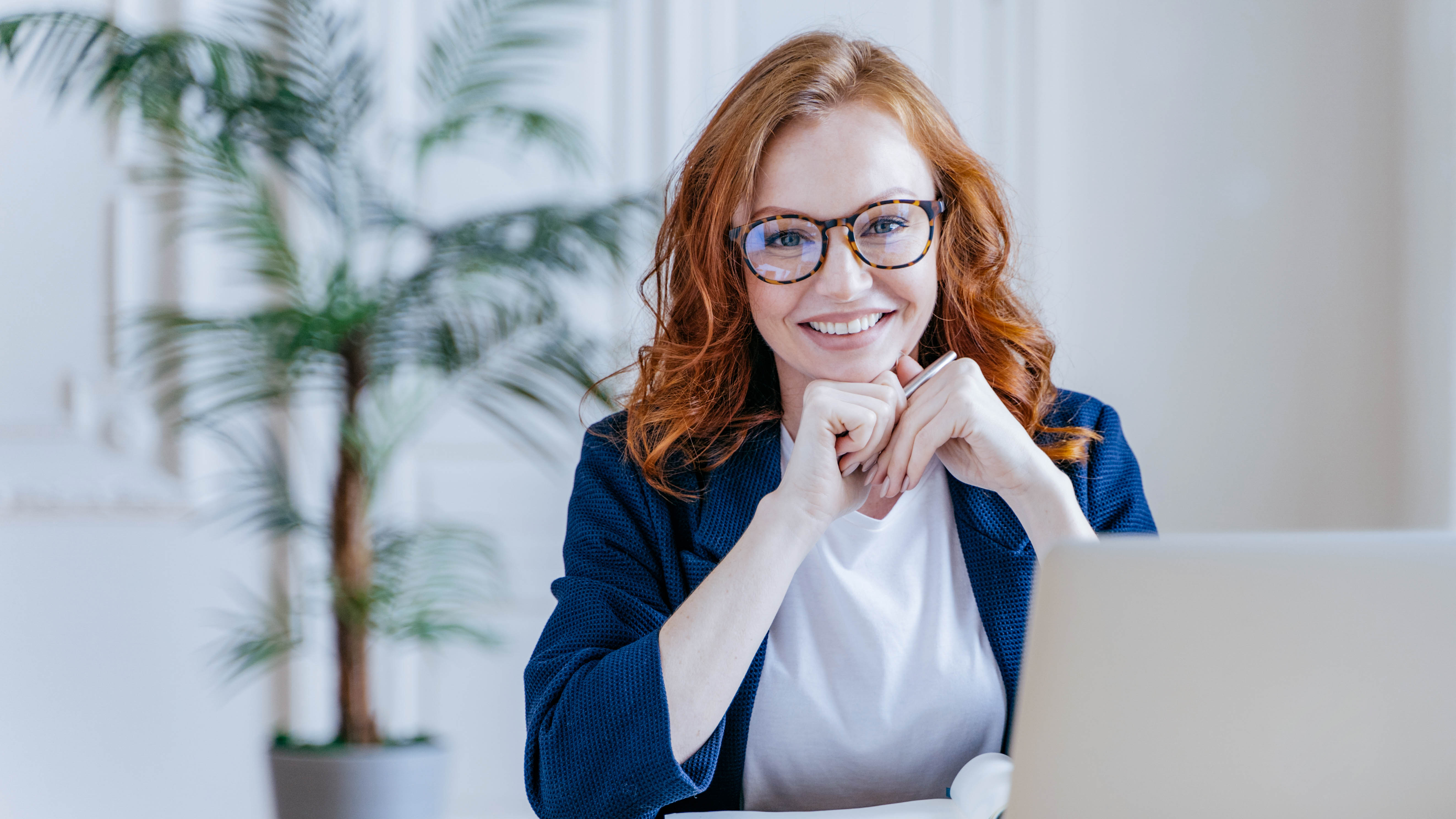 Lächelnde Frau mit roten Haaren und Brille sitzt am Schreibtisch vor einem Laptop, im Hintergrund eine Pflanze.