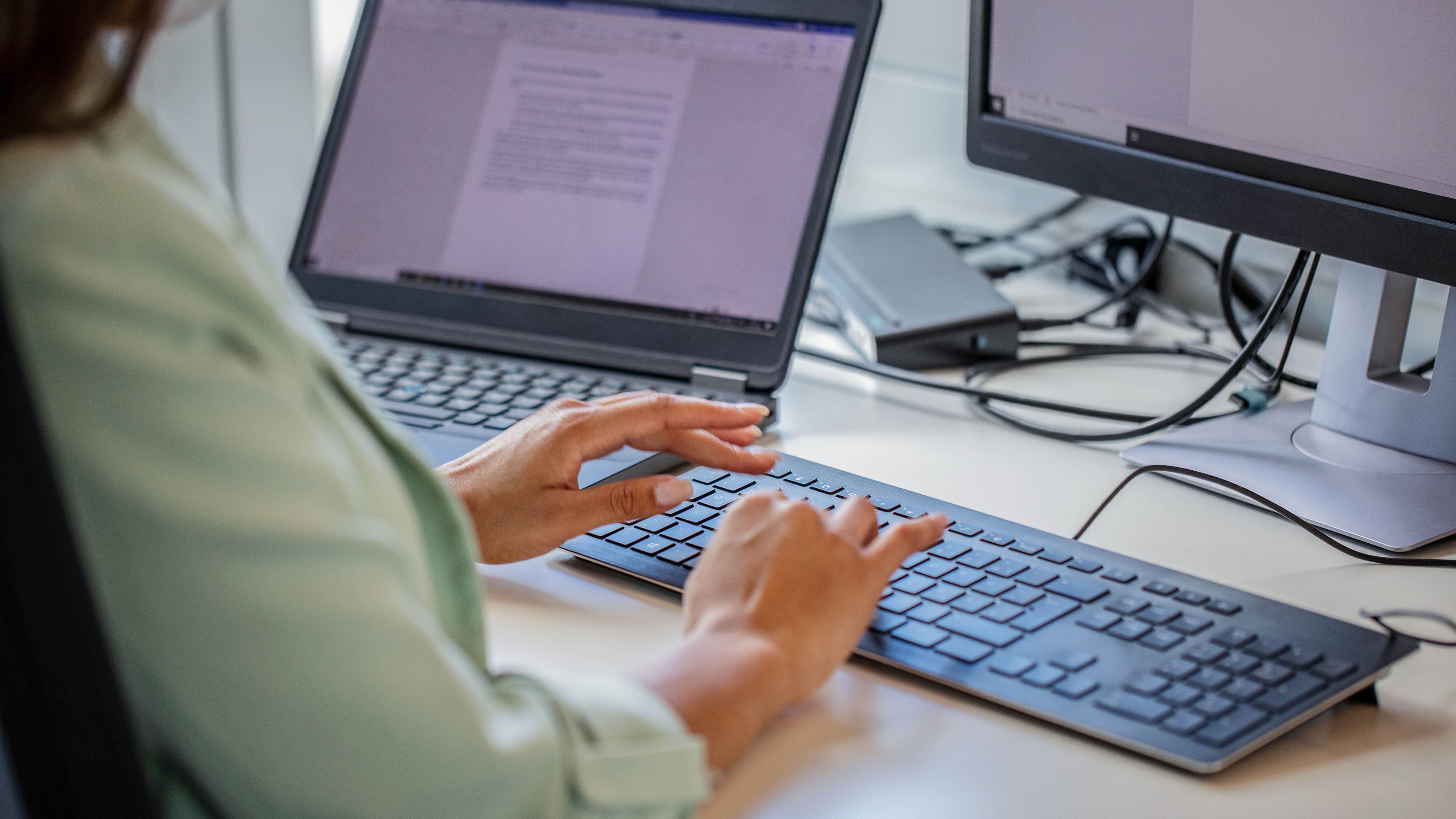 Hände tippen auf einer modernen Tastatur, während ein Laptop und ein Monitor im Hintergrund sichtbar sind.