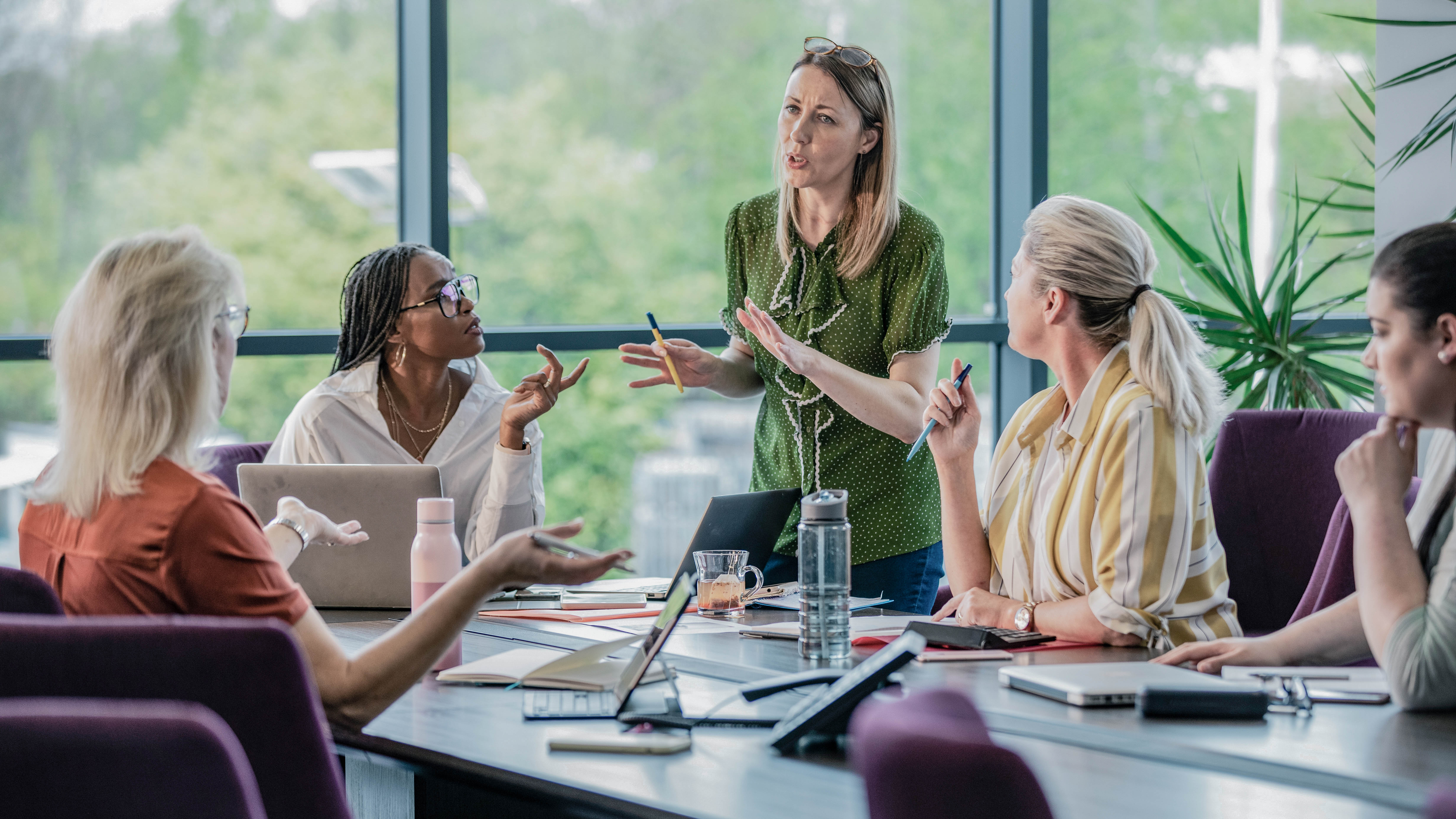 Eine Gruppe von fünf Frauen in einem modernen Konferenzraum diskutiert lebhaft, während Laptops und Notizen auf dem Tisch liegen.