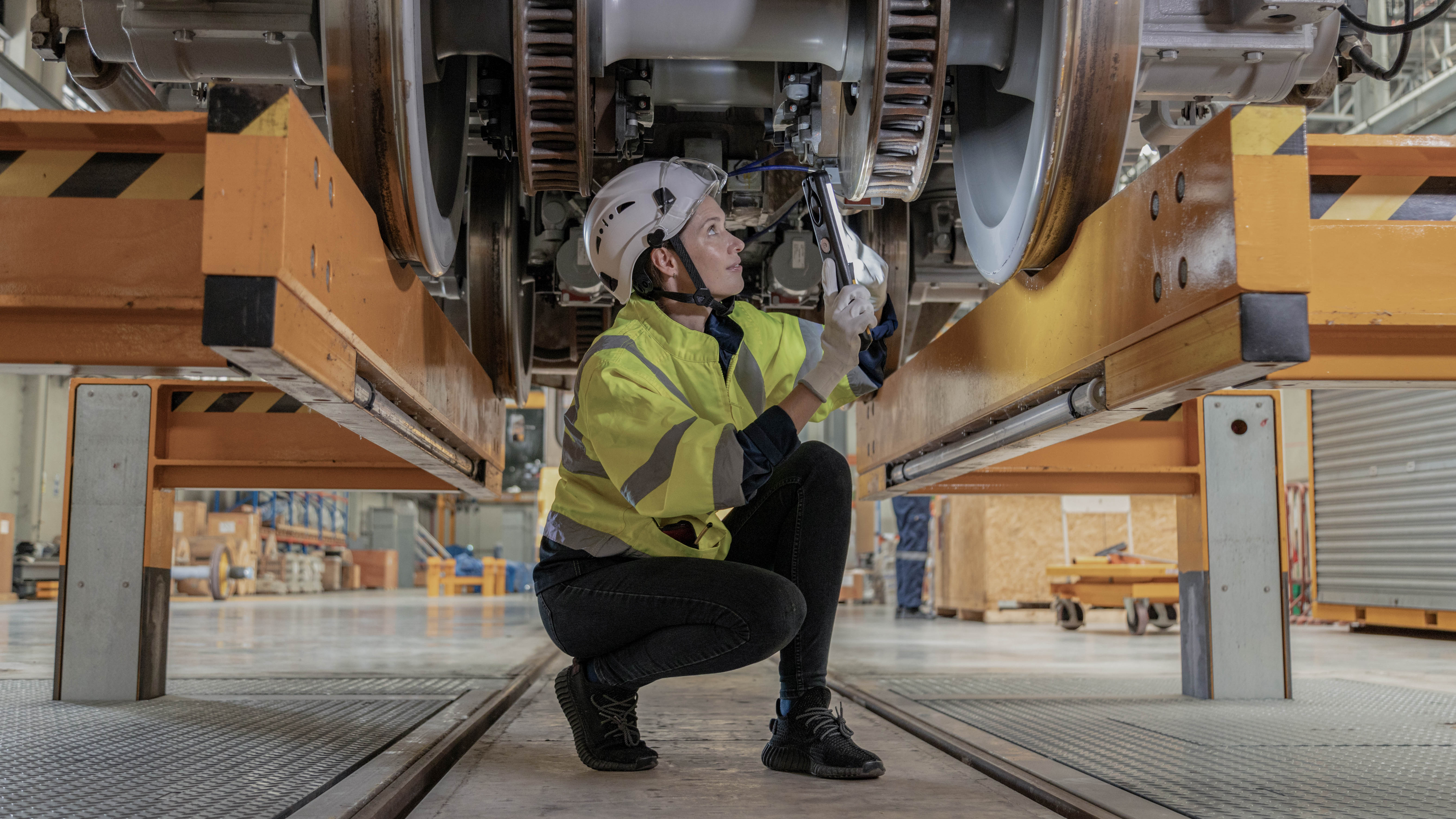 Eine Person in Sicherheitskleidung und Helm arbeitet mit einem Werkzeug unter einem großen Maschinenbauteil.