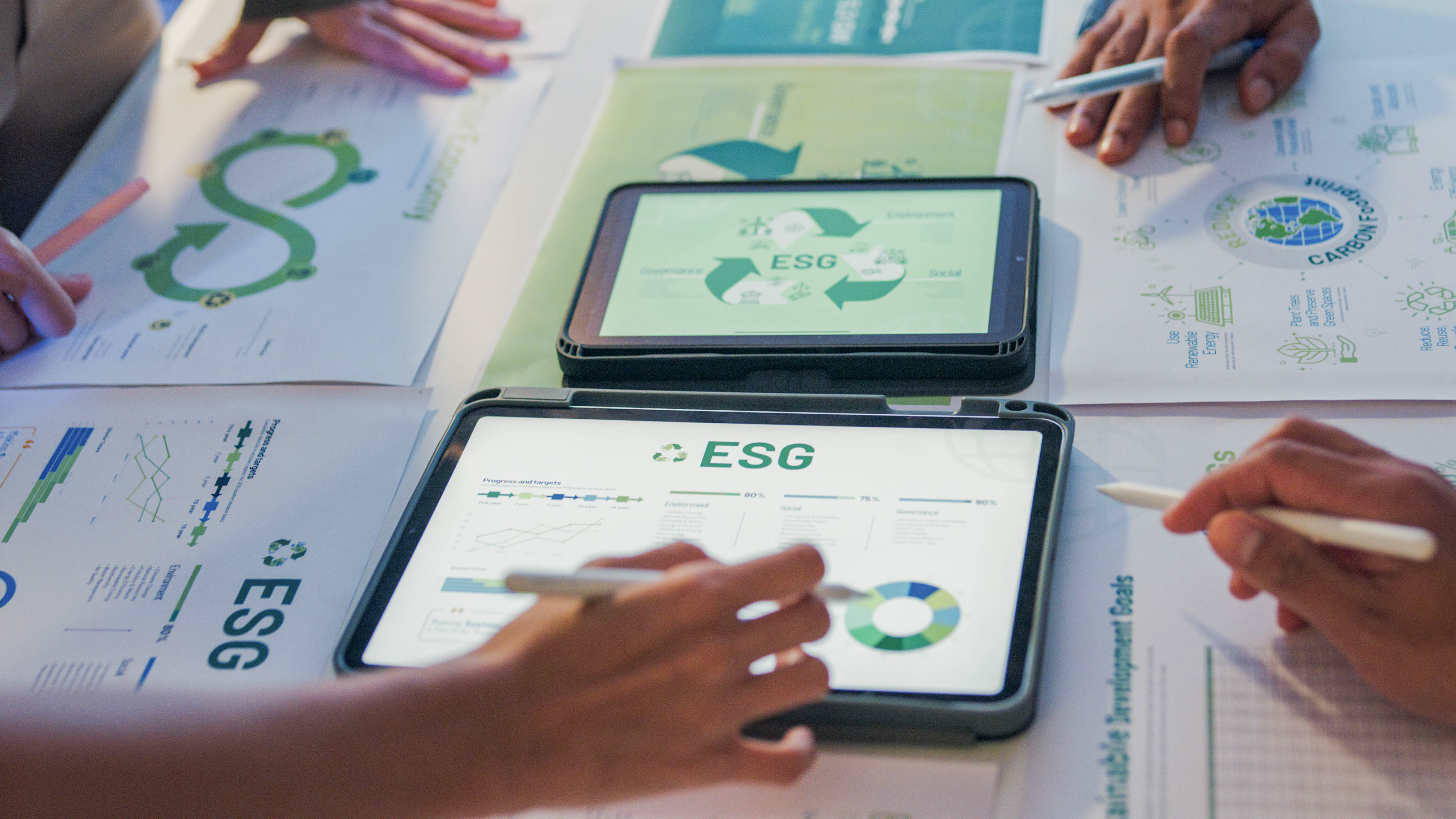 Hände und Tablets auf einem Tisch mit Dokumenten zu ESG-Themen und Diagrammen.