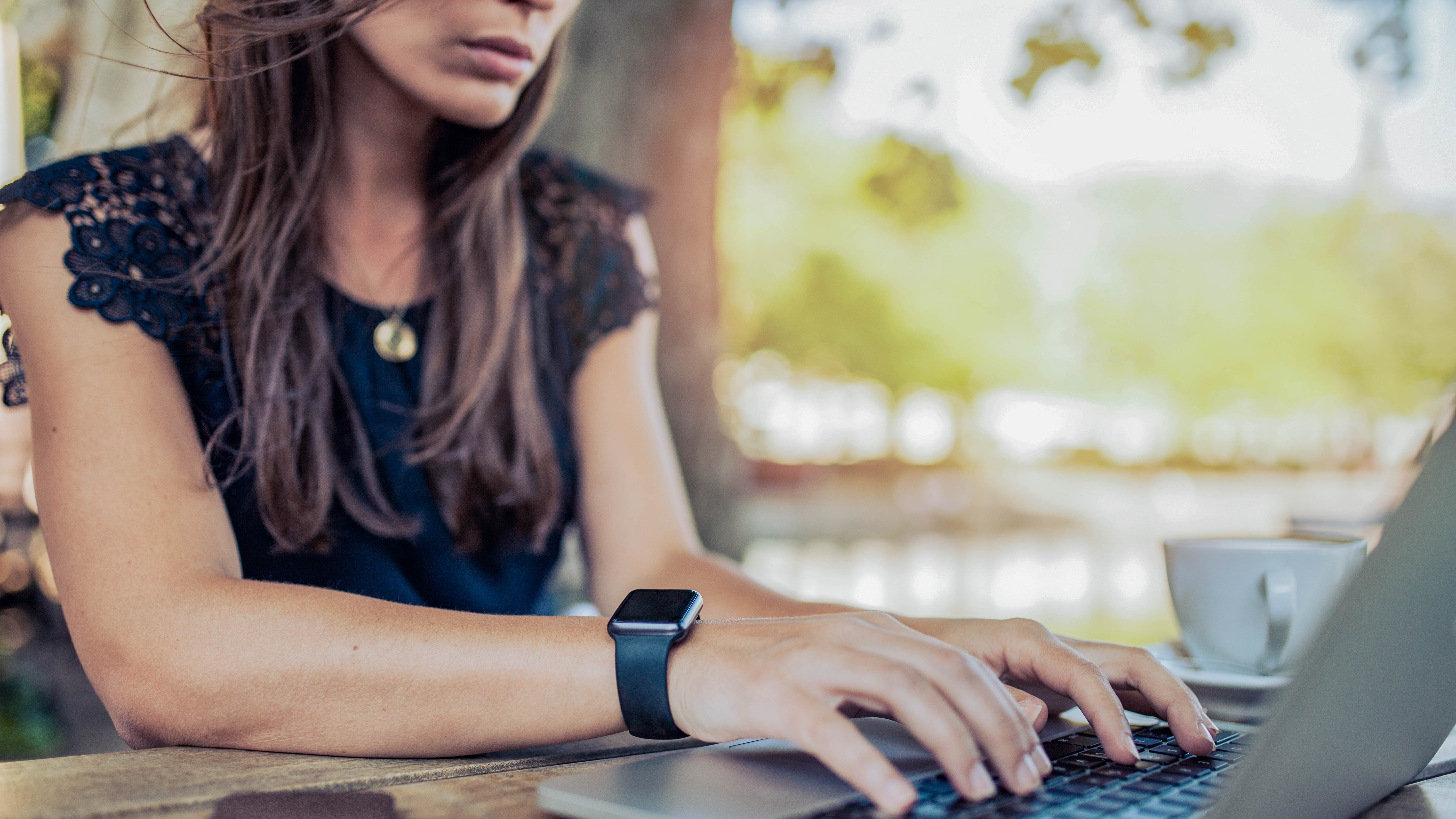 Eine Person mit langen Haaren tippt auf einem Laptop, während eine Tasse Kaffee neben ihr steht.