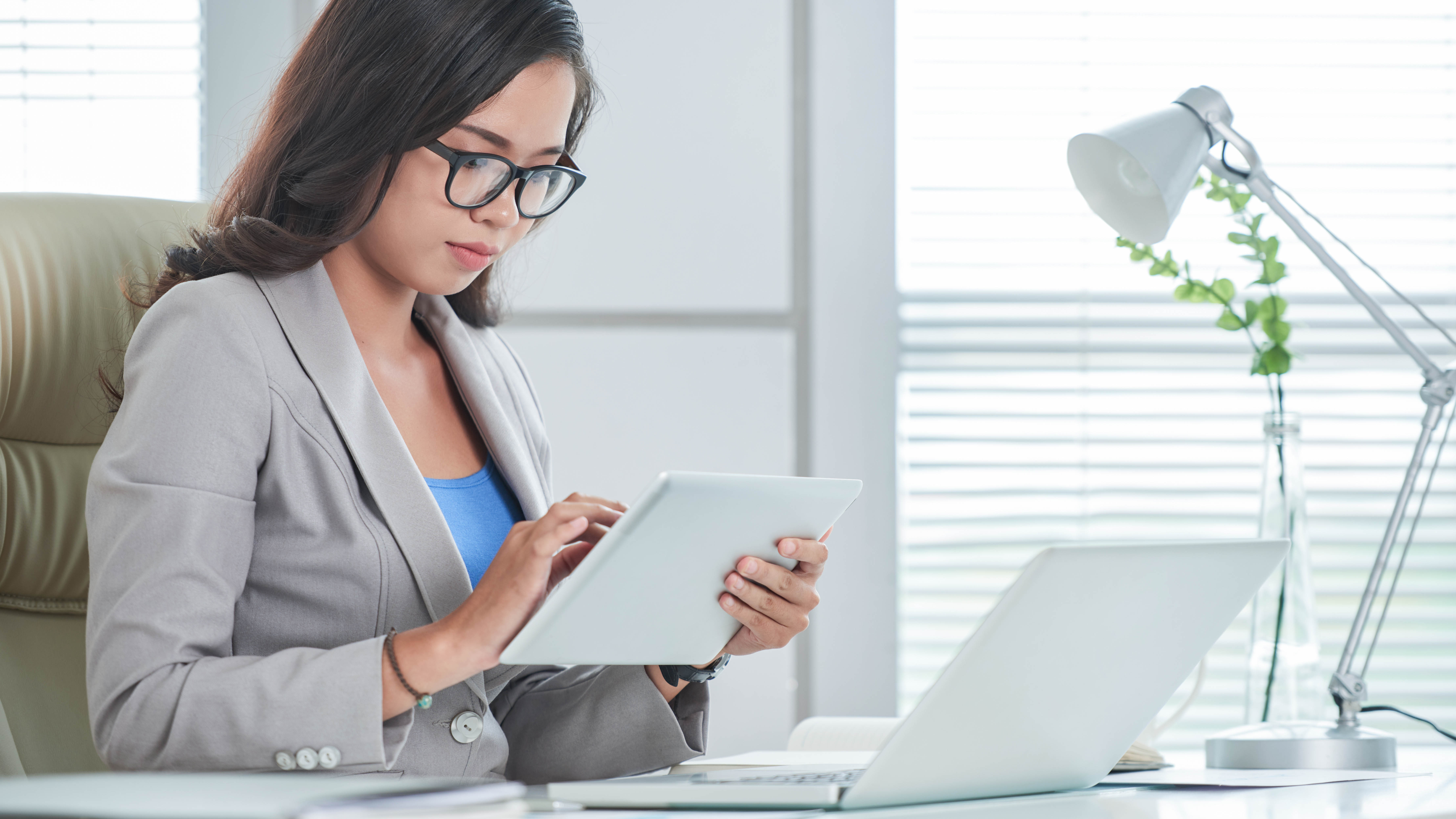 Eine Frau in einem grauen Blazer sitzt an einem Schreibtisch und nutzt ein Tablet.