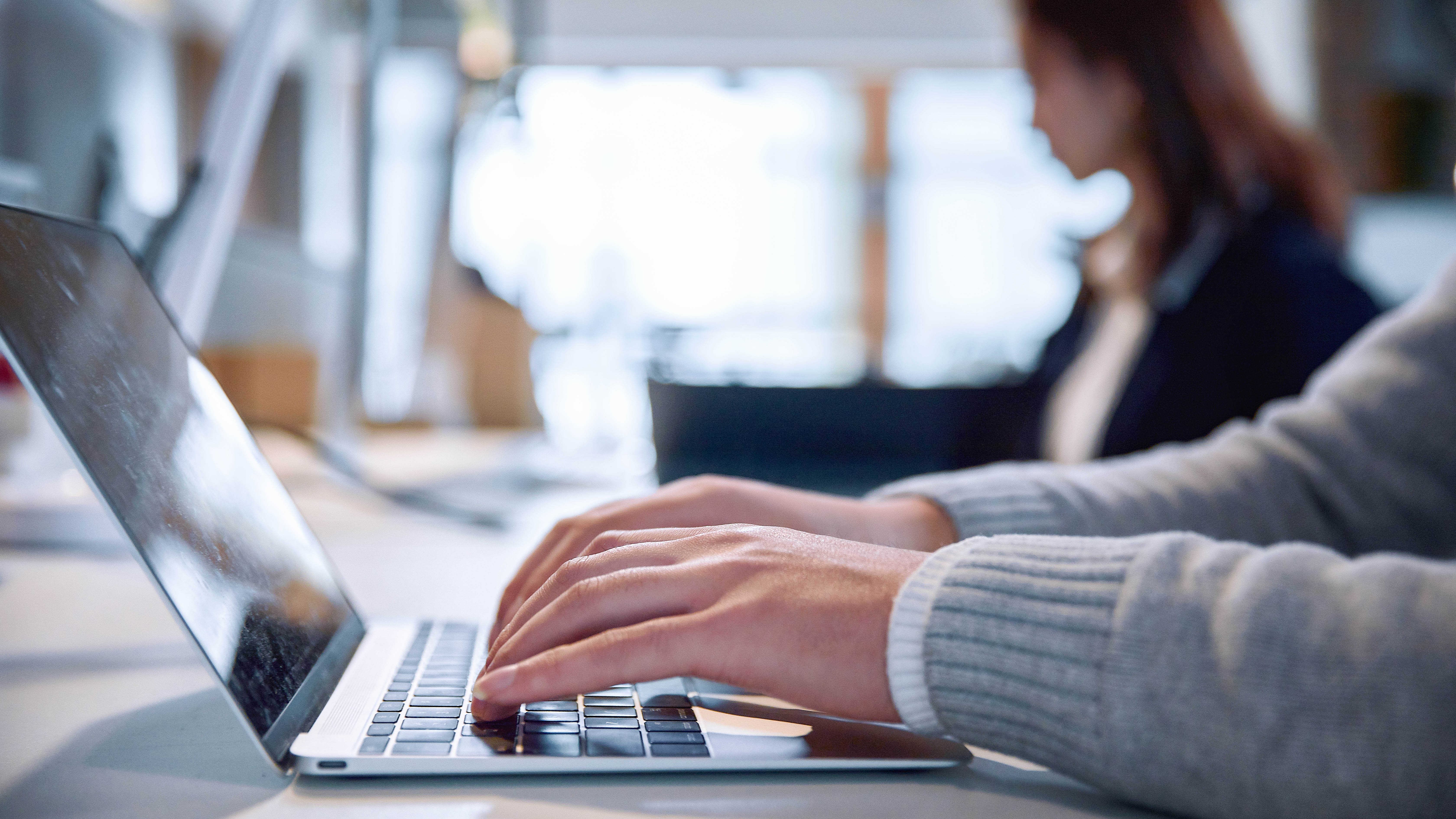 Hände tippen auf einer Laptop-Tastatur, im Hintergrund ist eine Person unscharf zu sehen.
