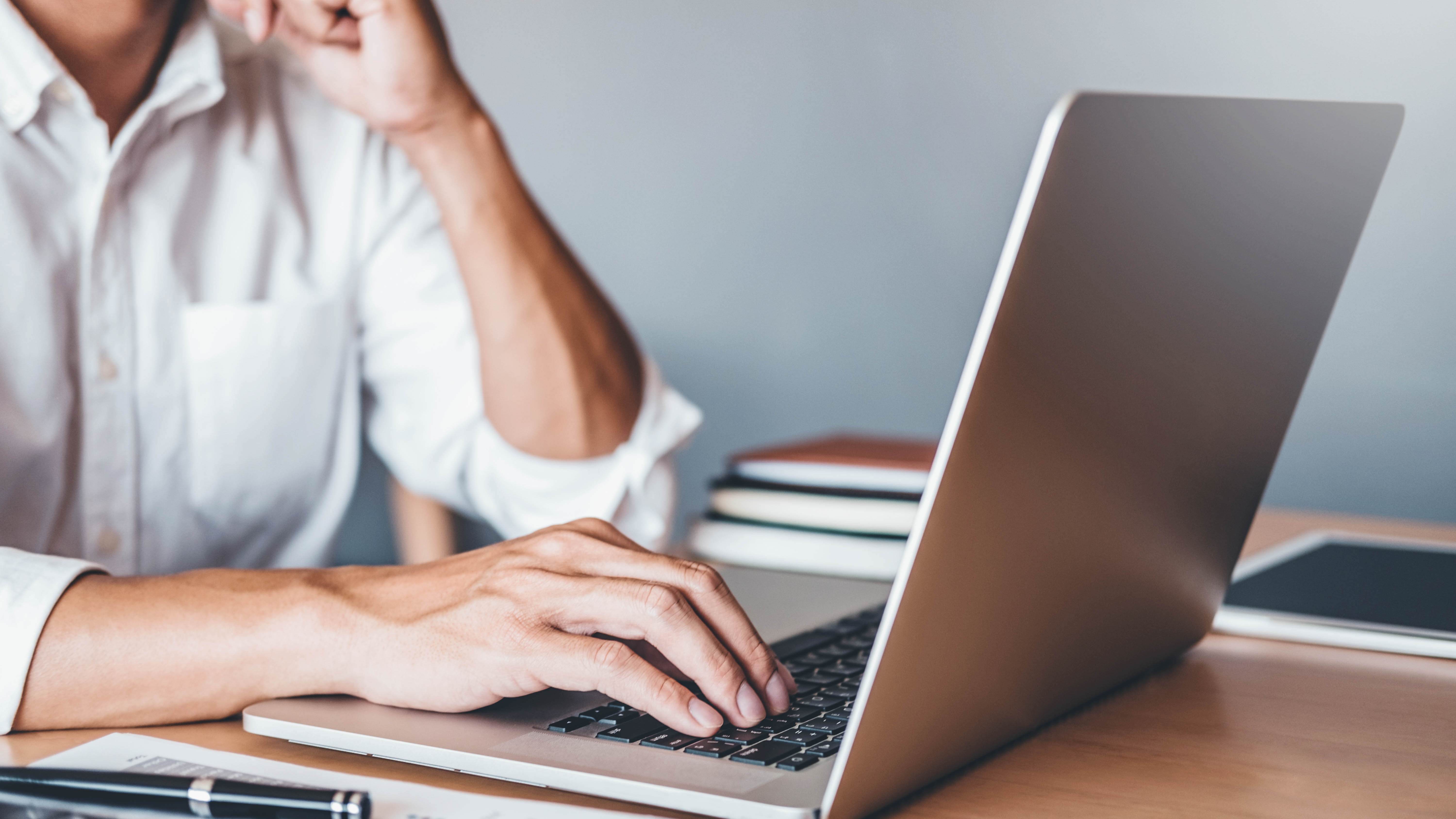 Eine Person tippt mit einer Hand auf einem Laptop, während die andere Hand das Kinn stützt.