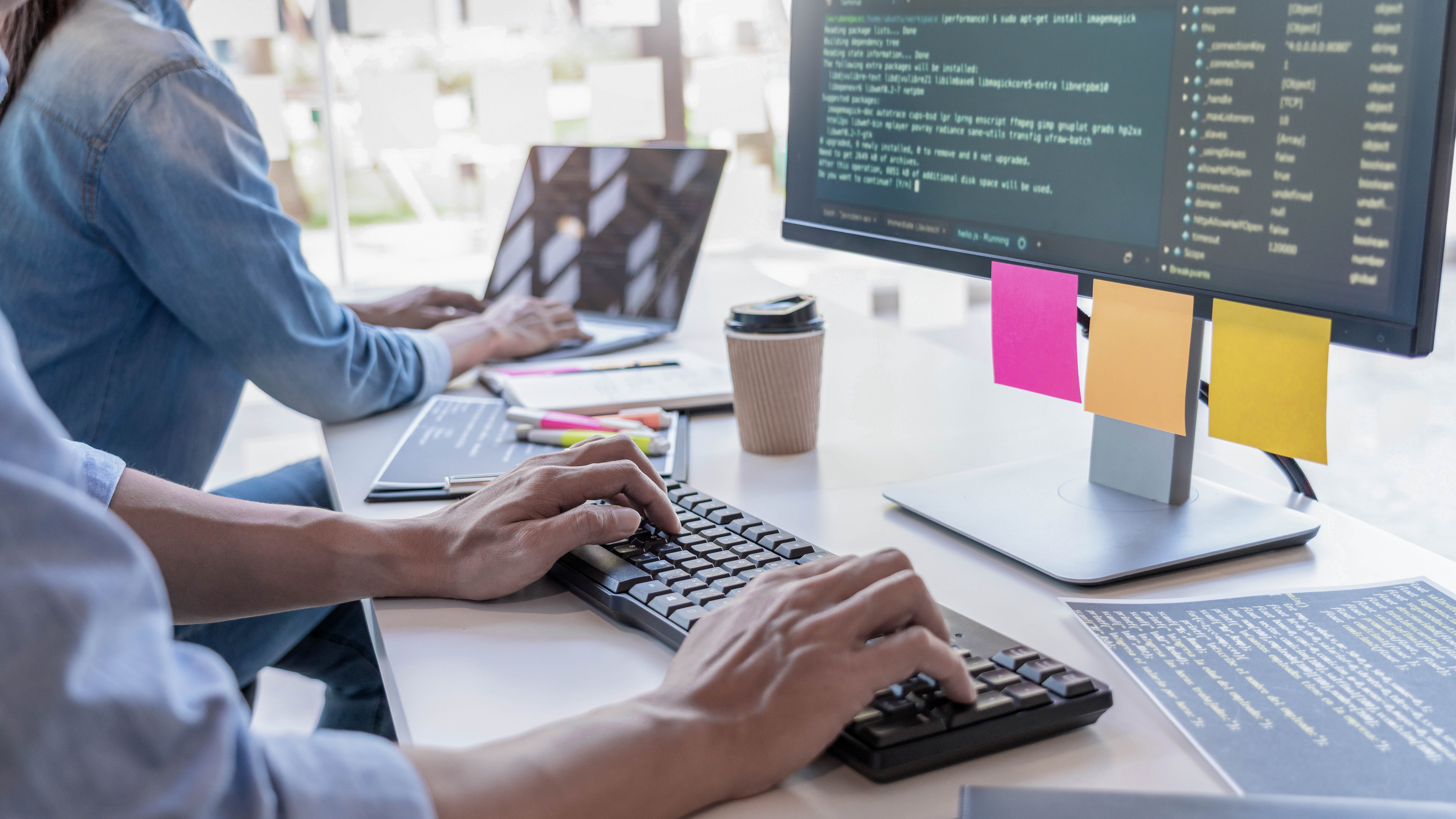 Hände tippen auf einer Tastatur, während ein Laptop und ein Monitor mit Programmiercode sichtbar sind.