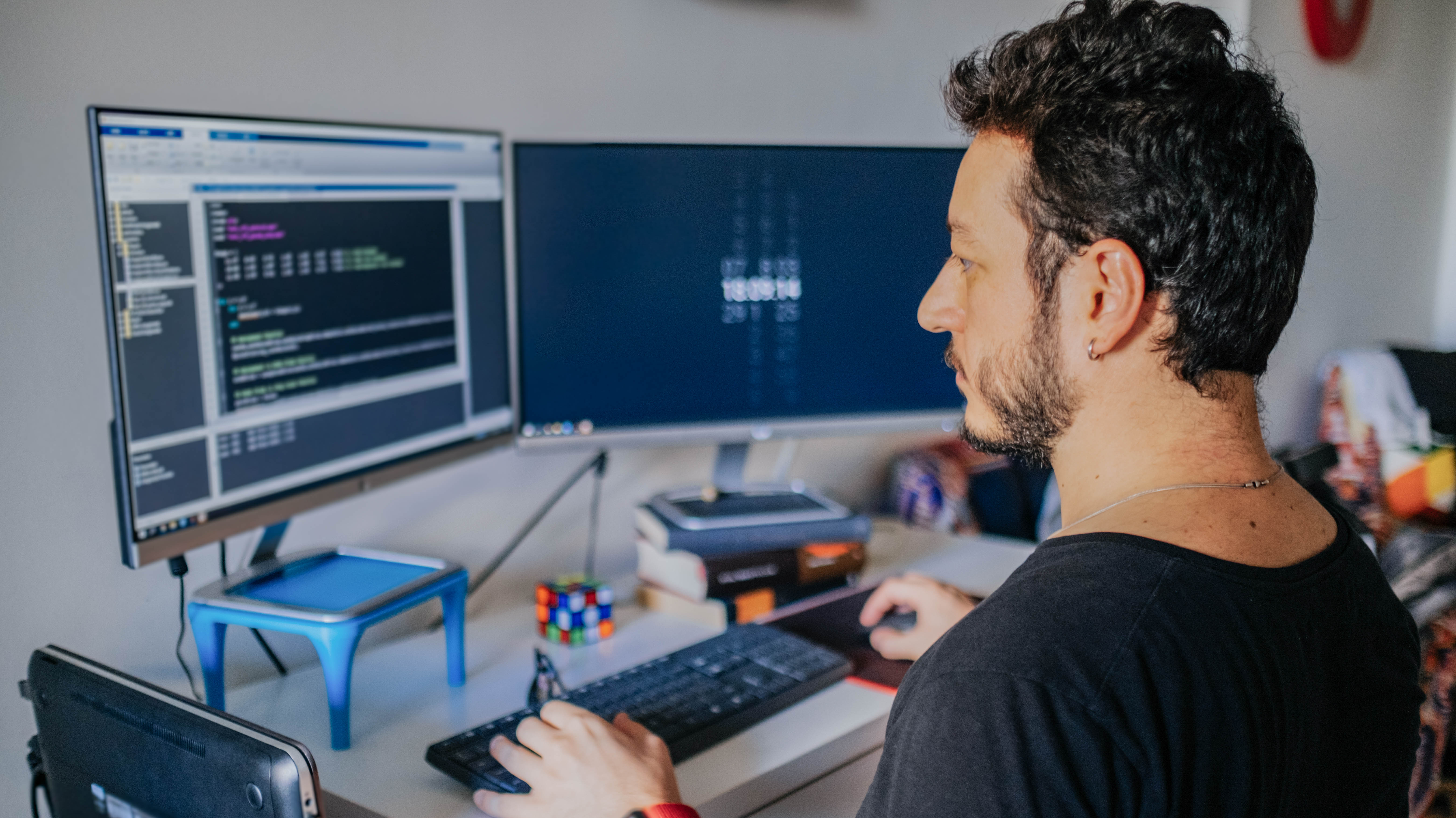 Mann sitzt an einem Schreibtisch mit zwei Monitoren, auf einem Bildschirm sind Programmierzeilen zu sehen.