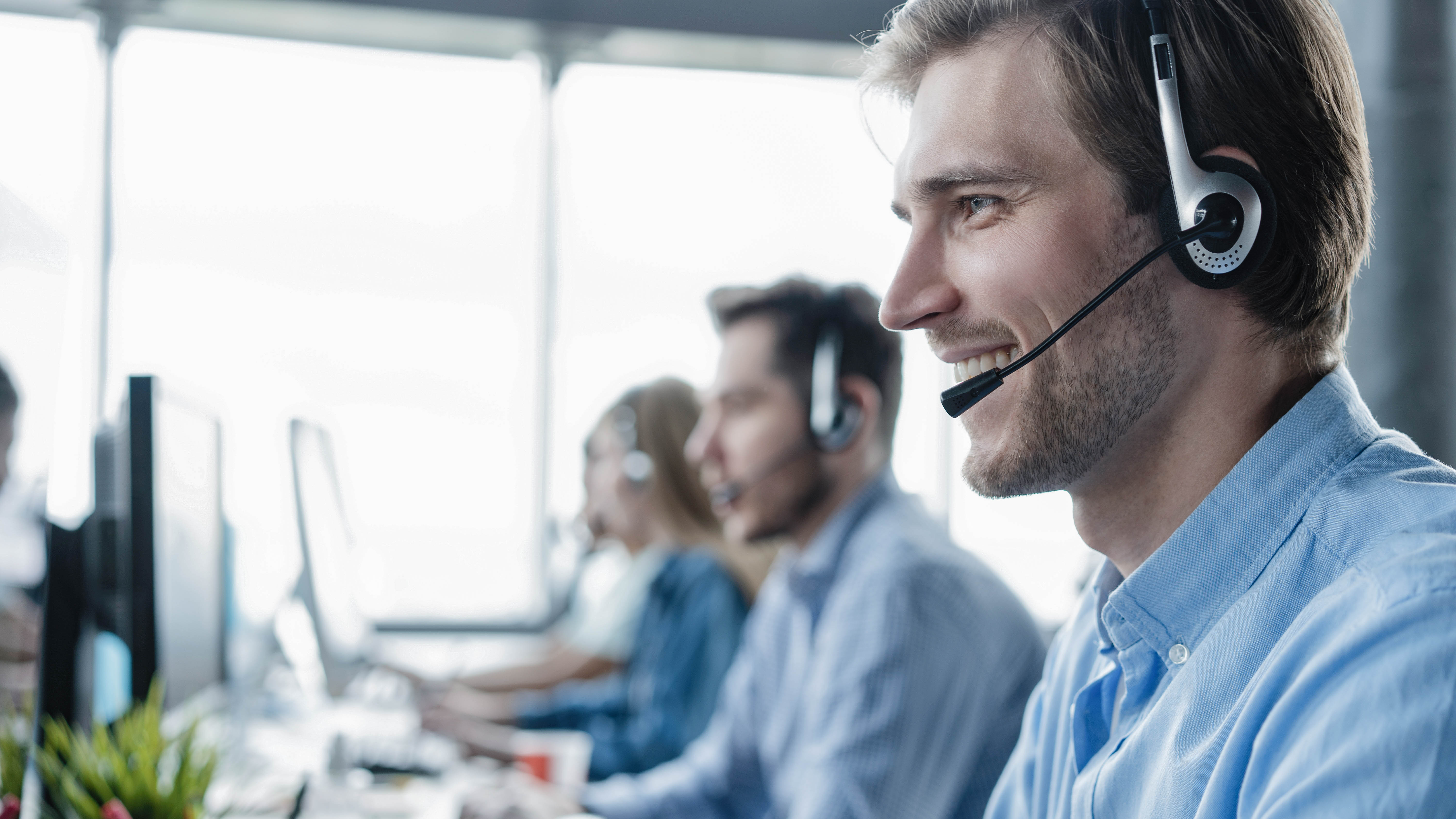 Mann mit Headset lächelt, während er an einem Computer arbeitet, im Hintergrund weitere Personen am Arbeitsplatz.