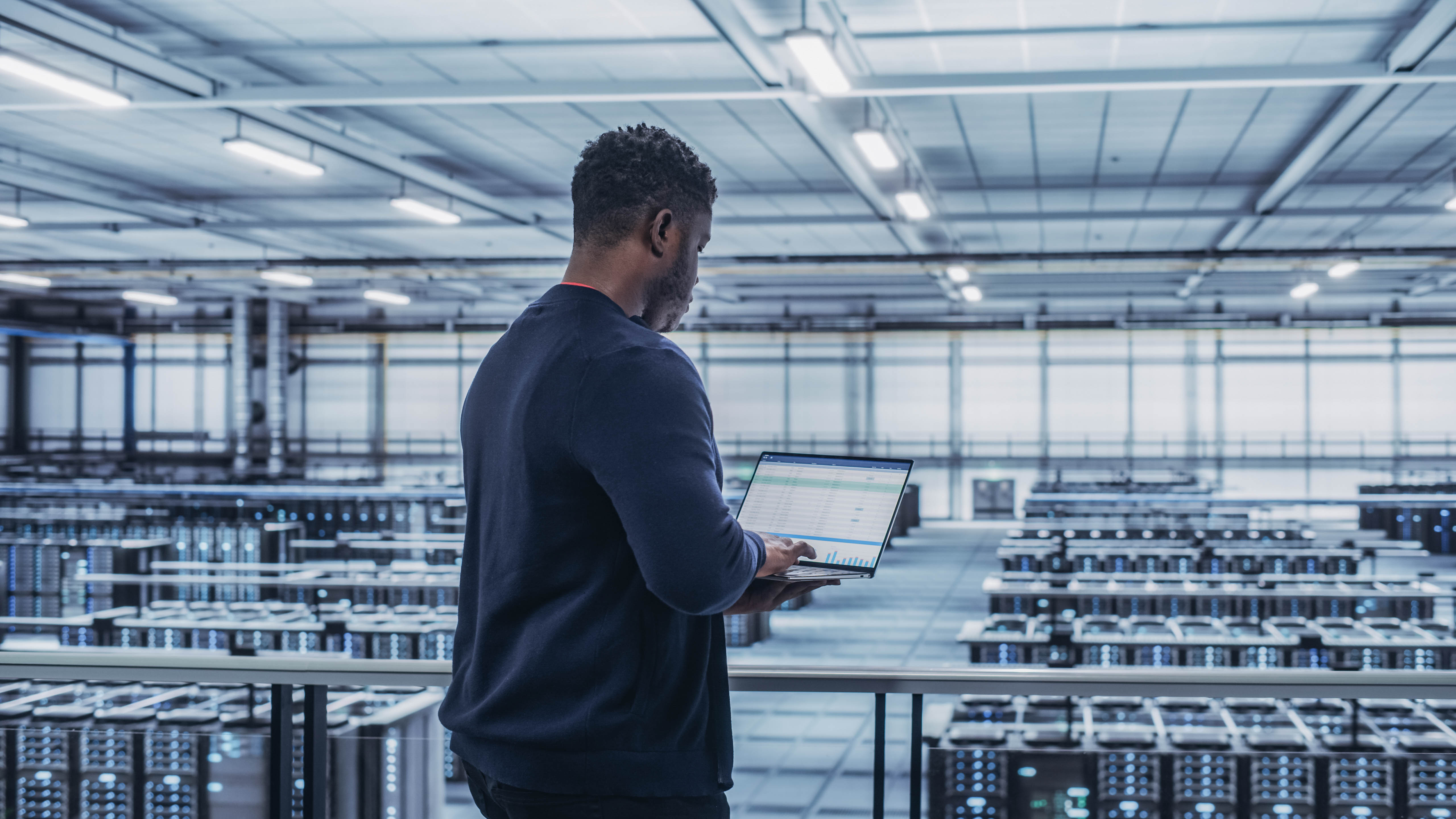 Mann mit Laptop steht in einem großen Raum voller Server-Racks und betrachtet Daten auf dem Bildschirm.