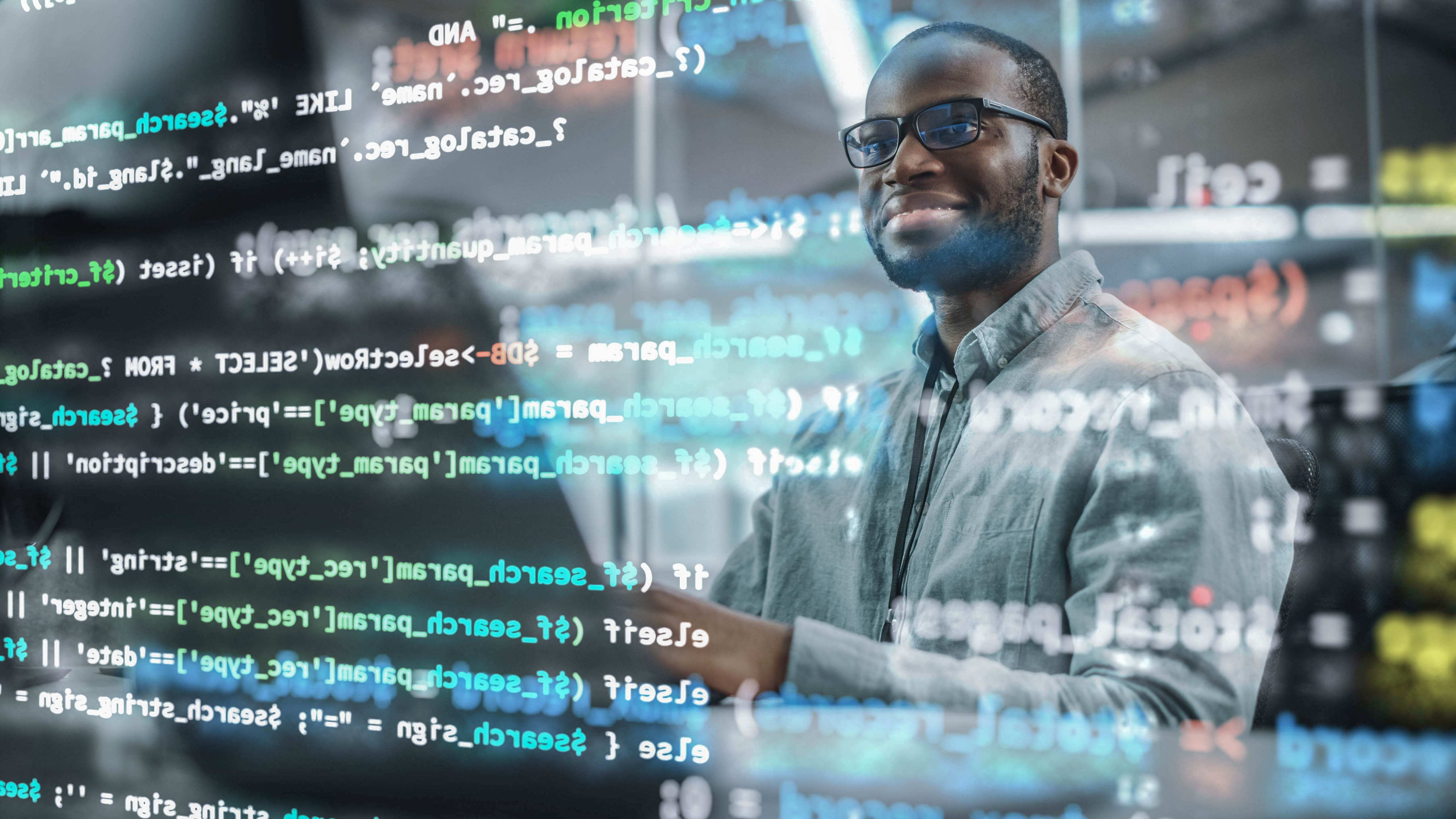Ein lächelnder Mann mit Brille arbeitet an einem Laptop, umgeben von Codezeilen auf einem Bildschirm.