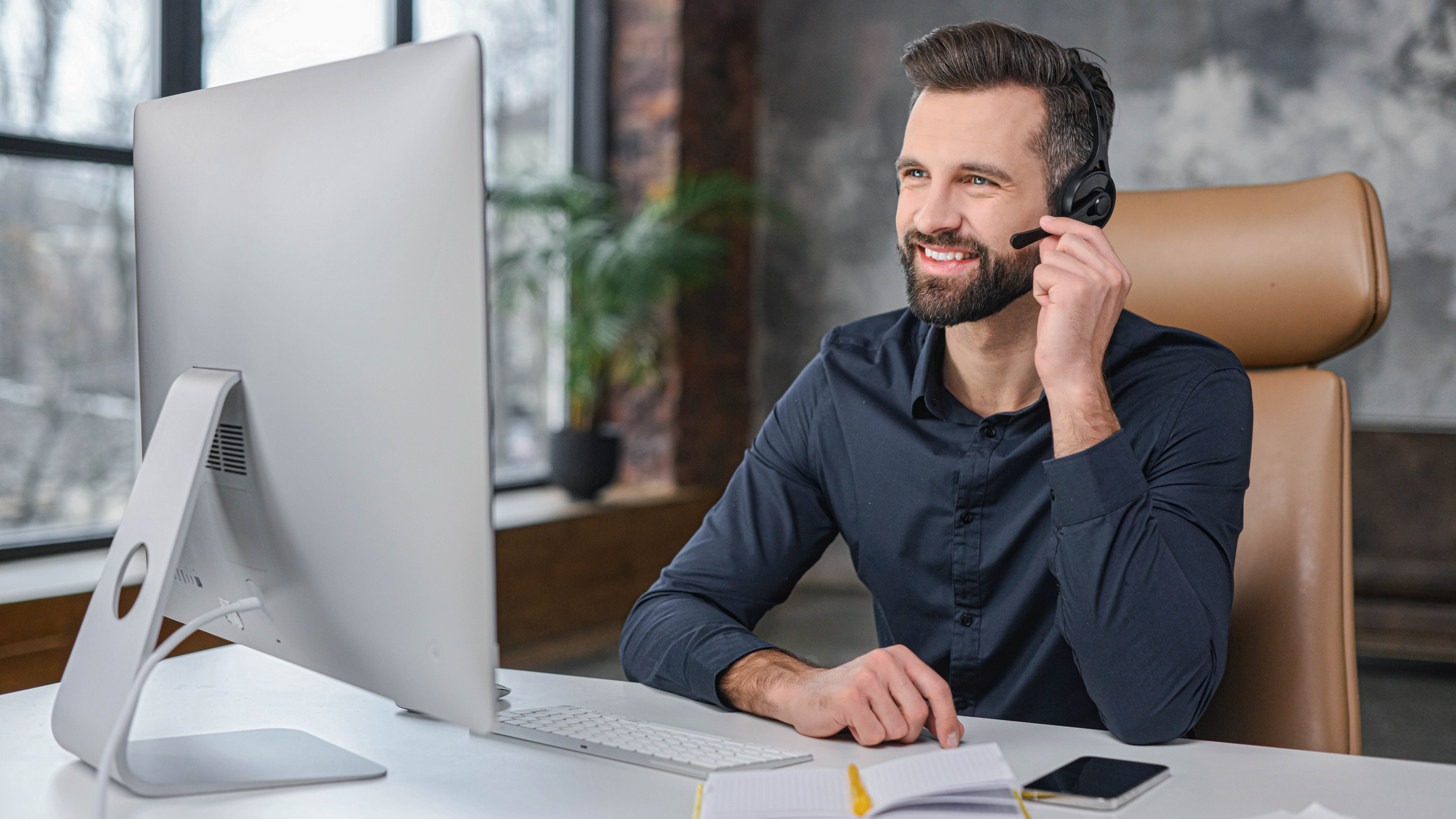 Mann mit Headset sitzt an einem Schreibtisch vor einem Computer und lächelt.