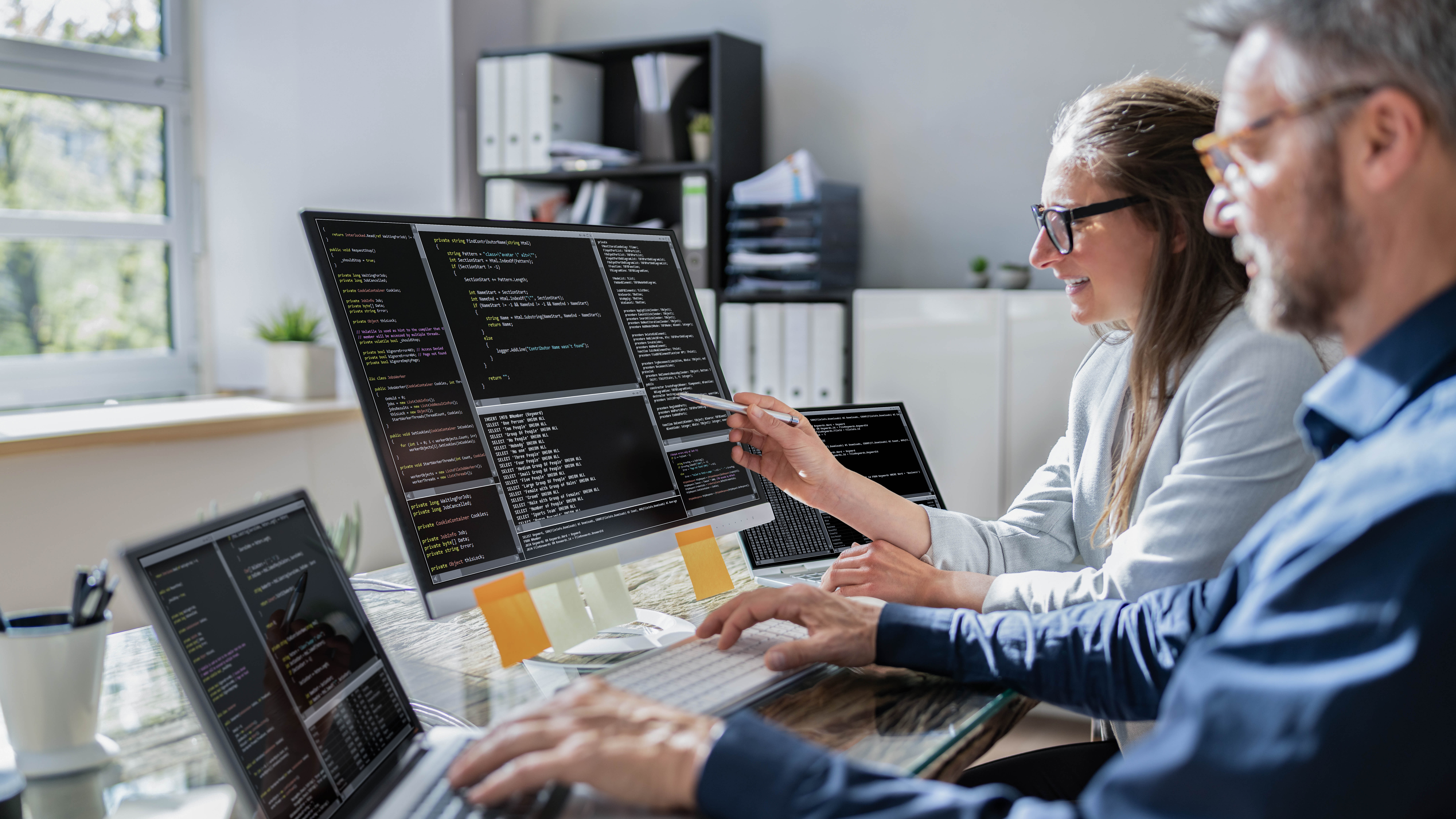 Zwei Personen arbeiten an mehreren Computern, die Programmiercode auf Bildschirmen anzeigen.