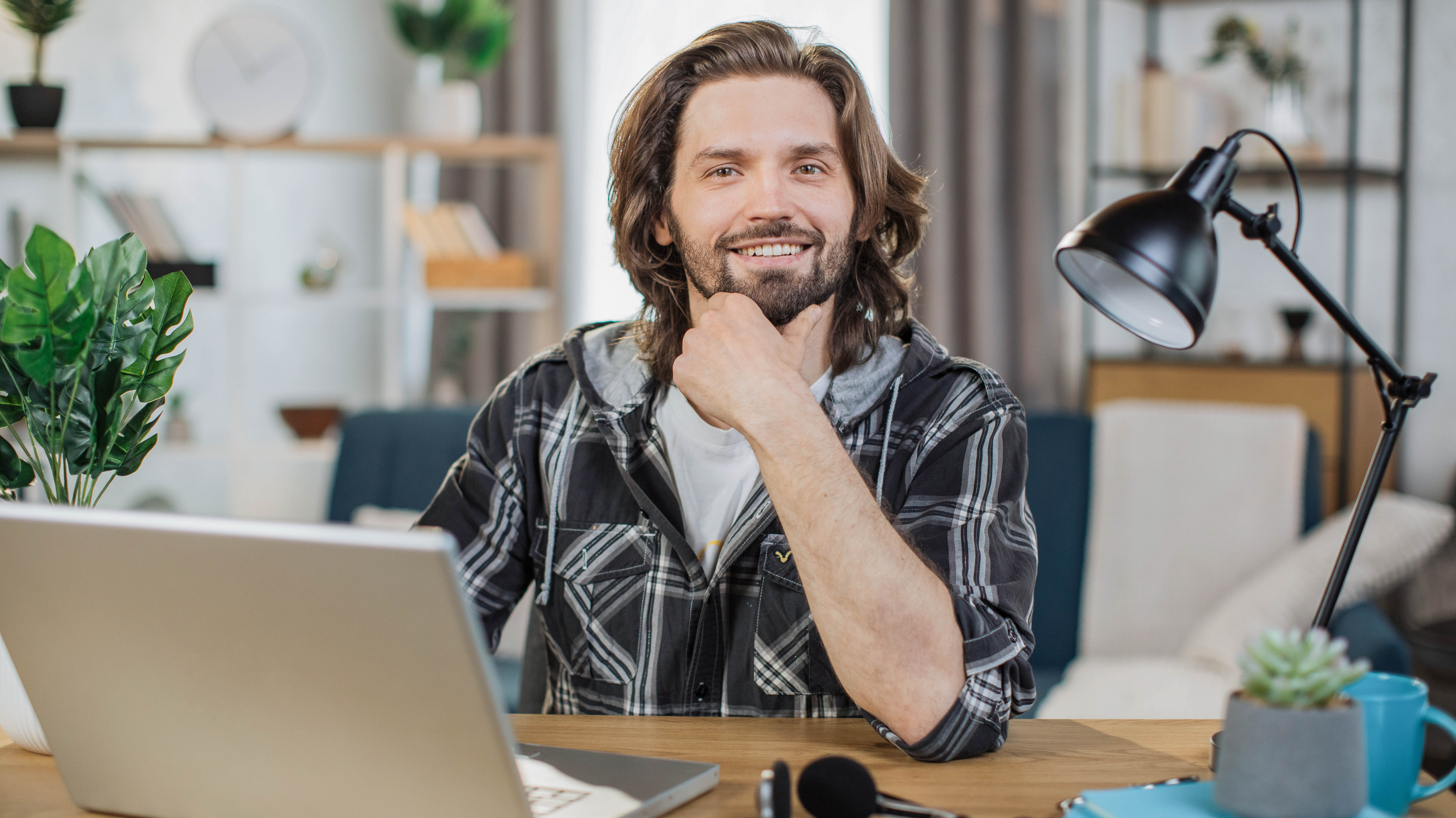 Lächelnder Mann mit langen Haaren sitzt an einem Tisch vor einem Laptop in einem modernen Raum.
