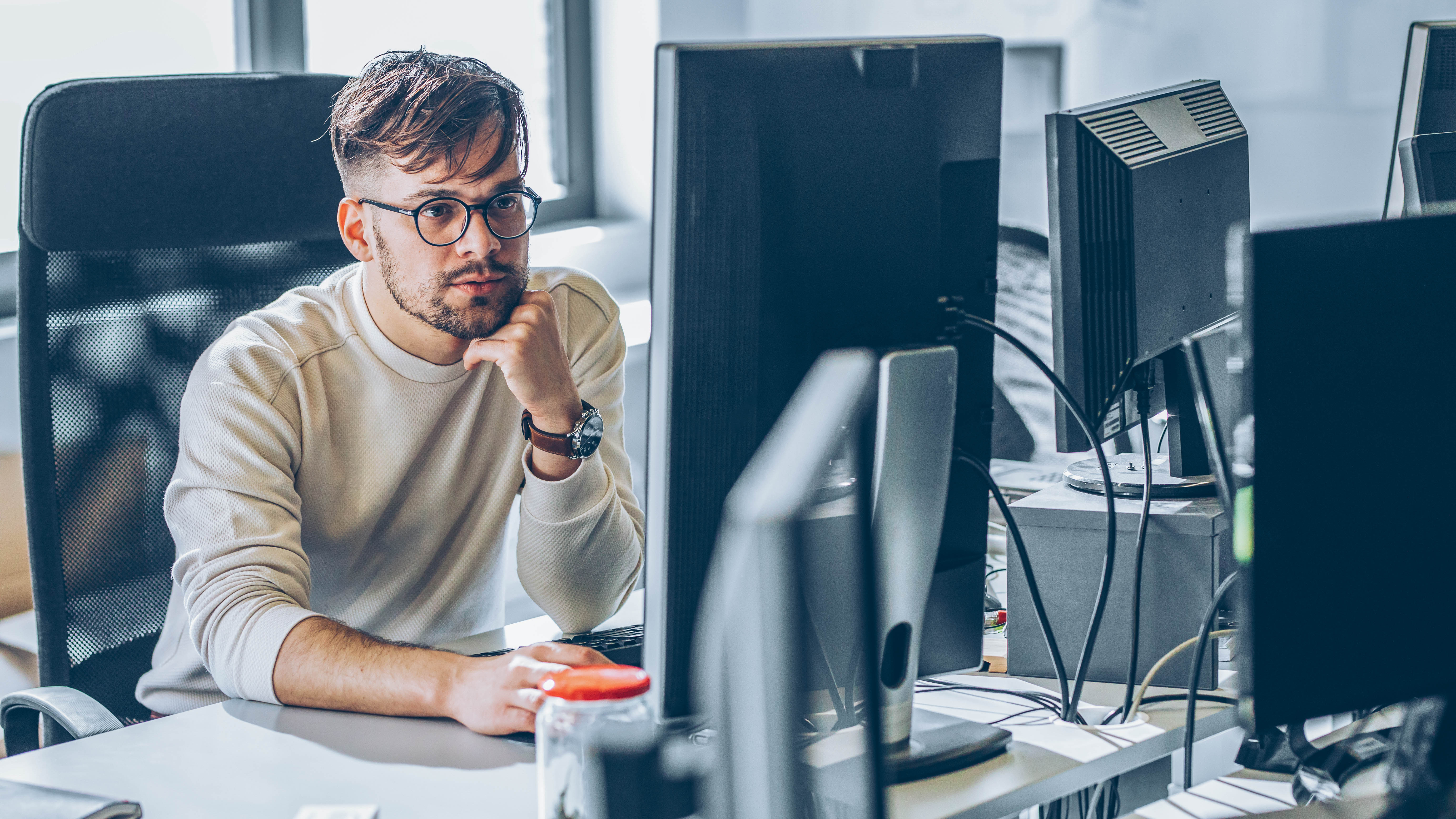 Ein junger Mann mit Brille sitzt nachdenklich vor einem Computerbildschirm in einem modernen Büro.