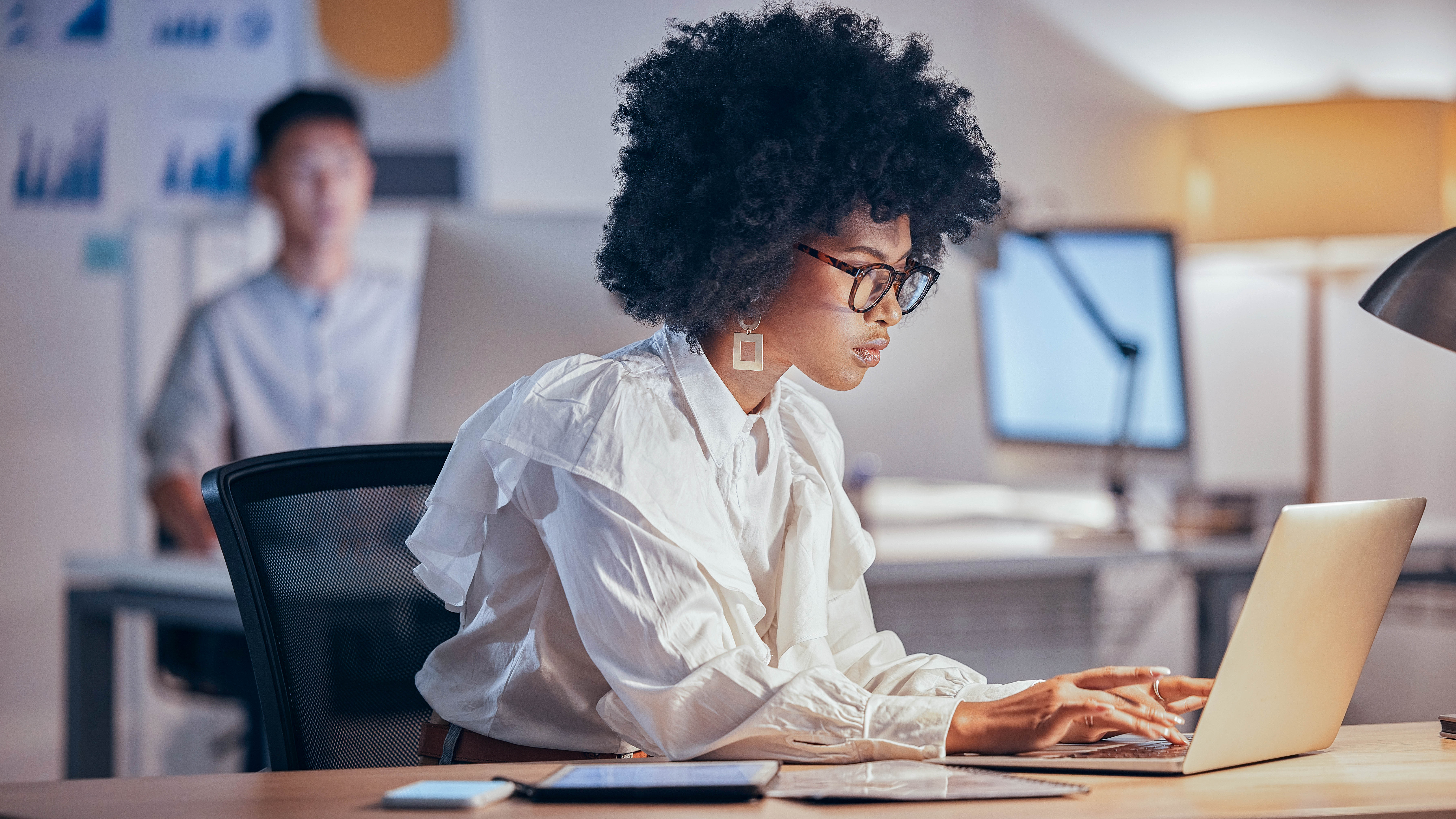 Eine Person mit lockigem Haar und Brille arbeitet konzentriert an einem Laptop in einem modernen Büro.
