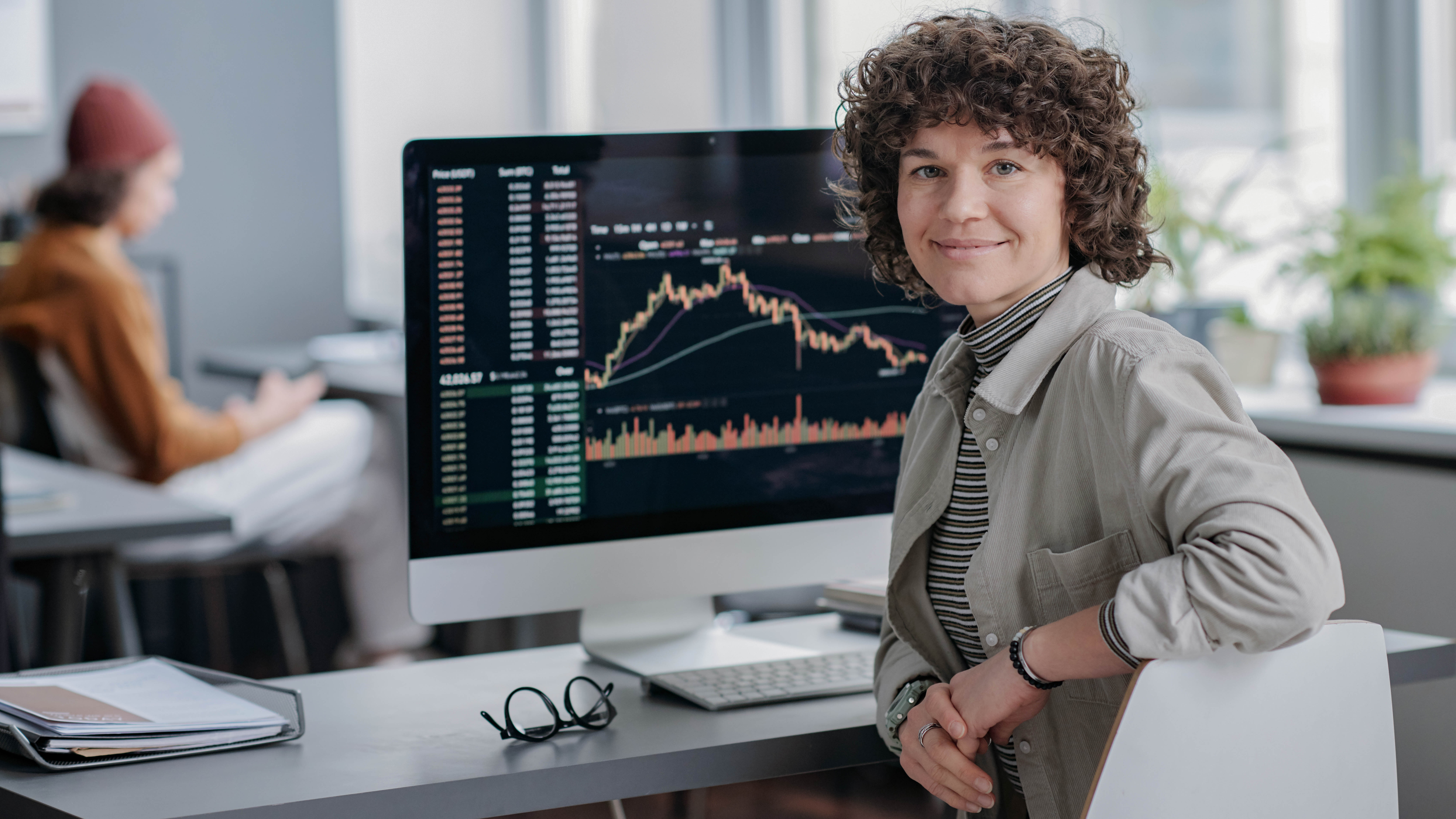 Eine Person mit lockigem Haar sitzt an einem Schreibtisch vor einem Computer mit Finanzdiagrammen.