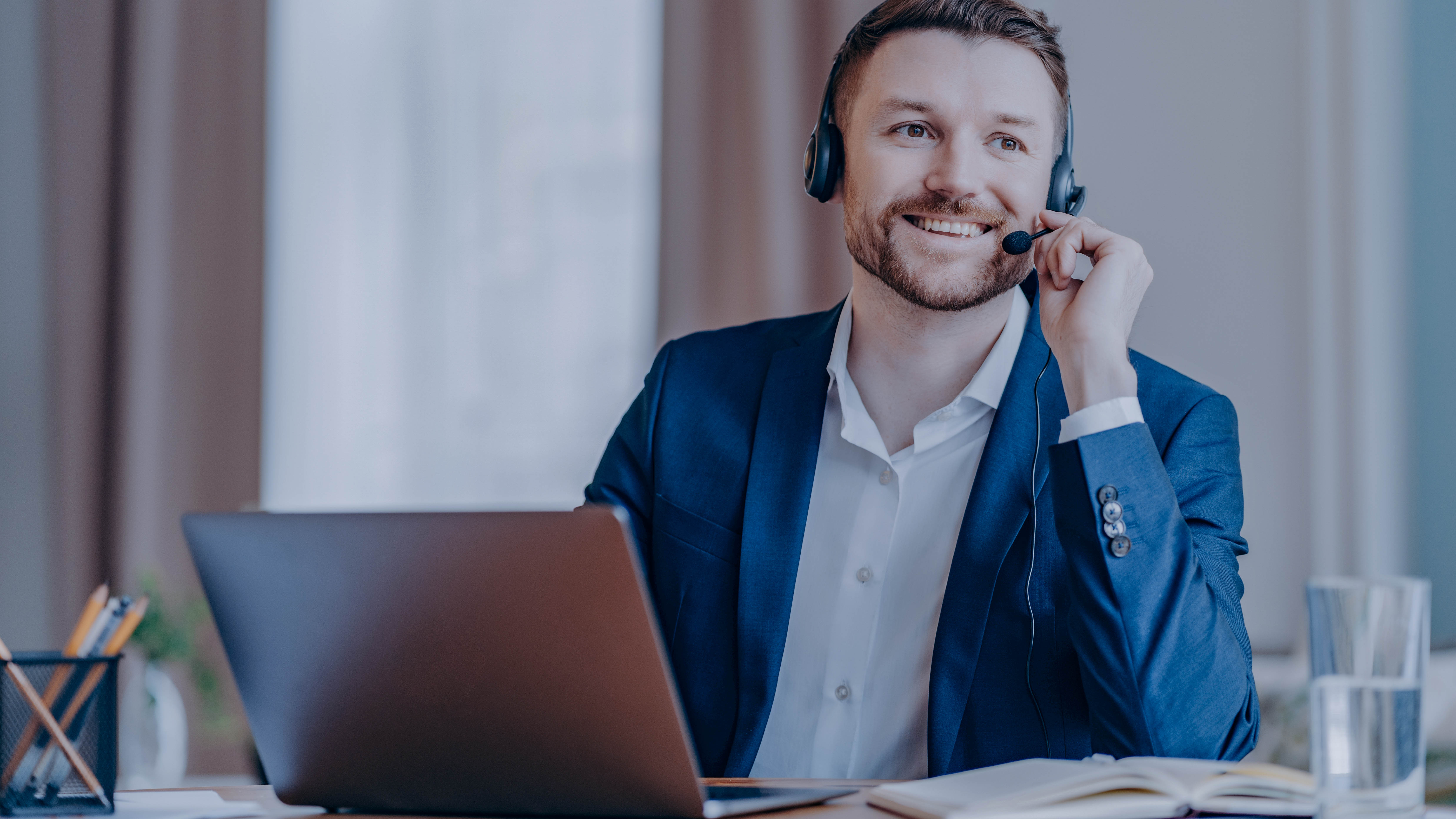 Mann mit Headset sitzt an einem Schreibtisch mit Laptop, lächelt und spricht in ein Mikrofon.