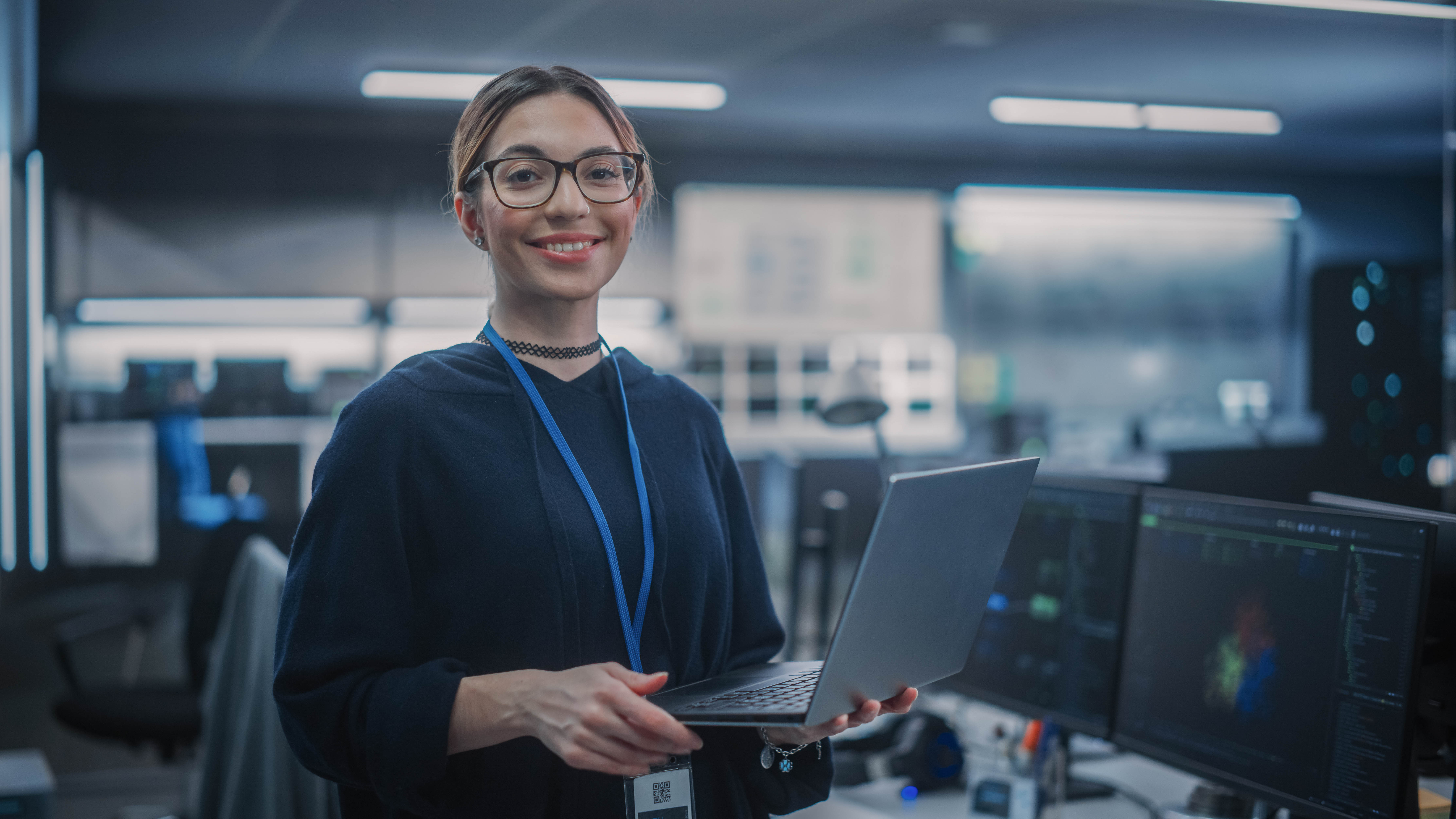 Frau mit Brille hält einen Laptop in einem modernen Büro mit Computerarbeitsplätzen im Hintergrund.