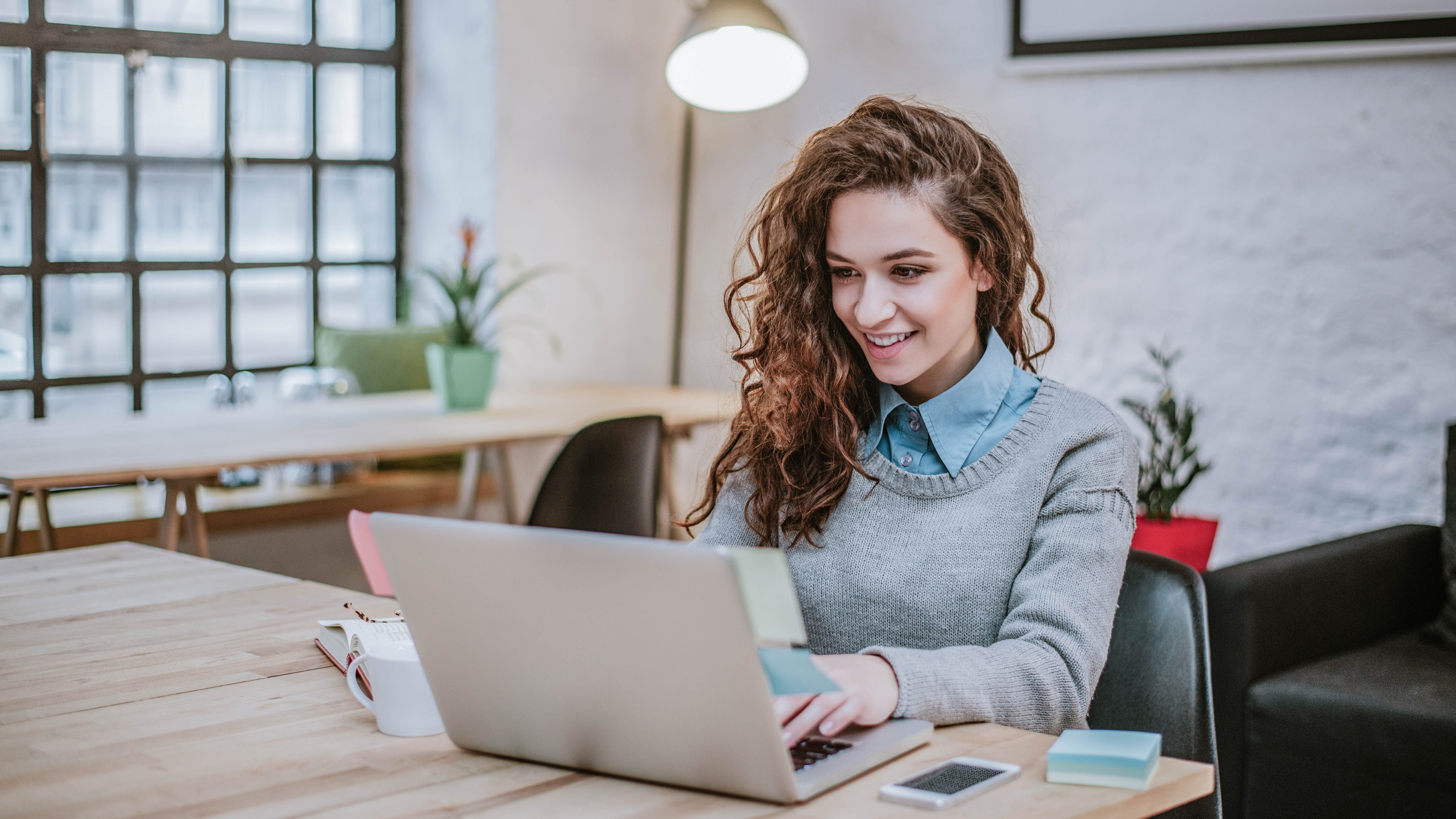 Junge Frau mit lockigem Haar sitzt an einem Tisch und arbeitet an einem Laptop in einem modernen Café.