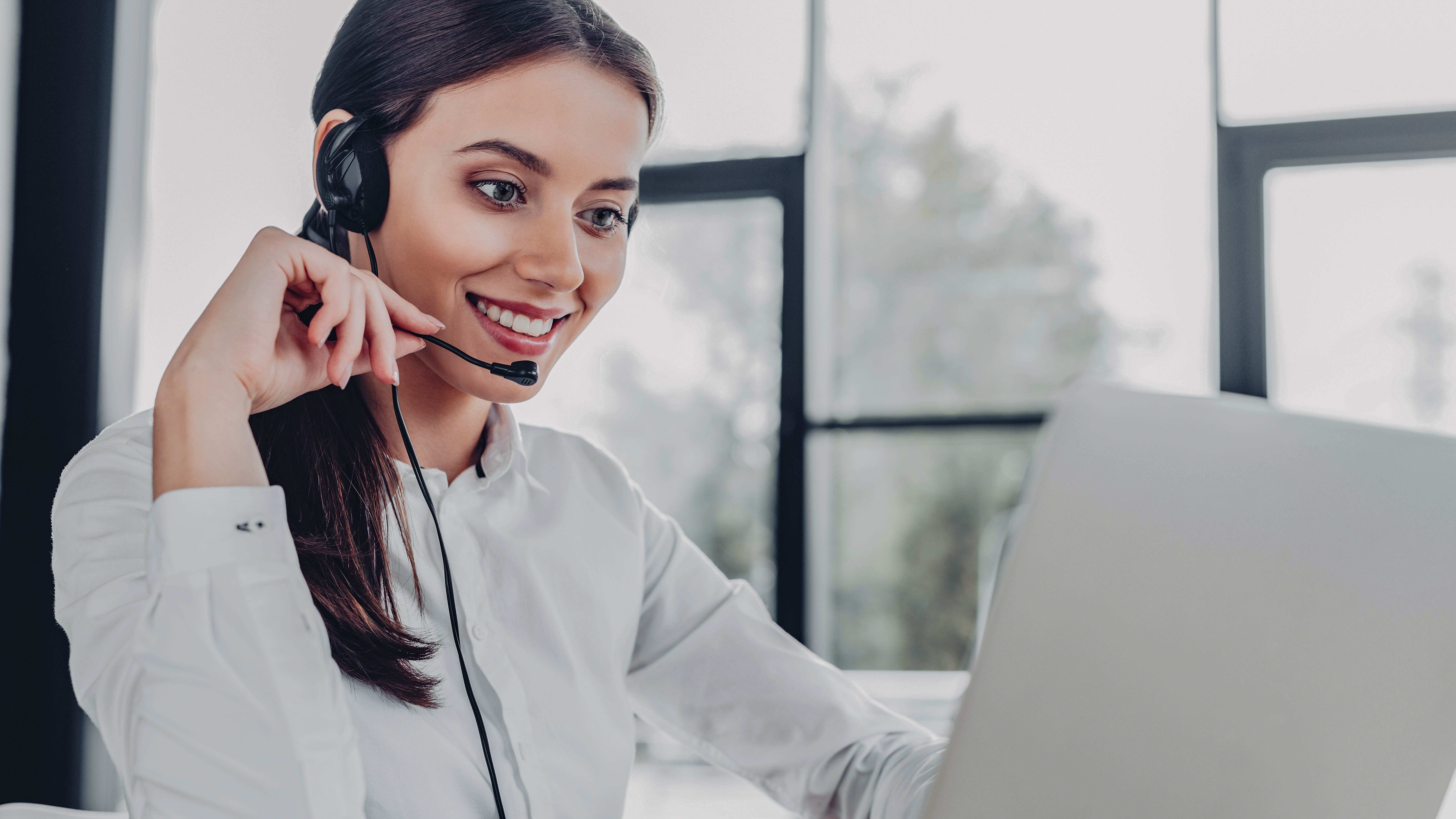 Frau mit Headset lächelt, während sie an einem Laptop arbeitet, im Hintergrund große Fenster.