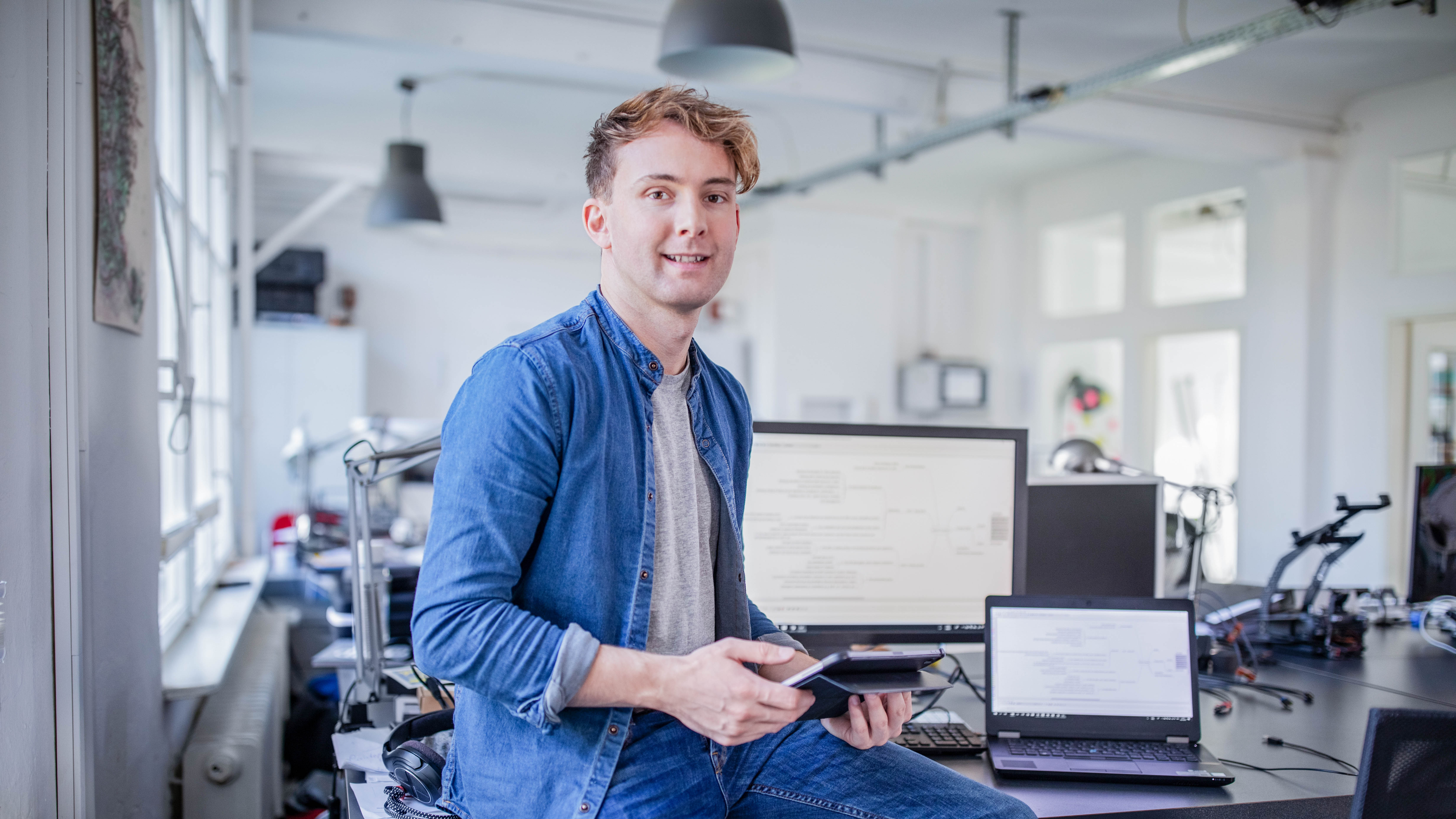Junger Mann sitzt an einem Schreibtisch mit mehreren Computern und hält ein Tablet in der Hand.